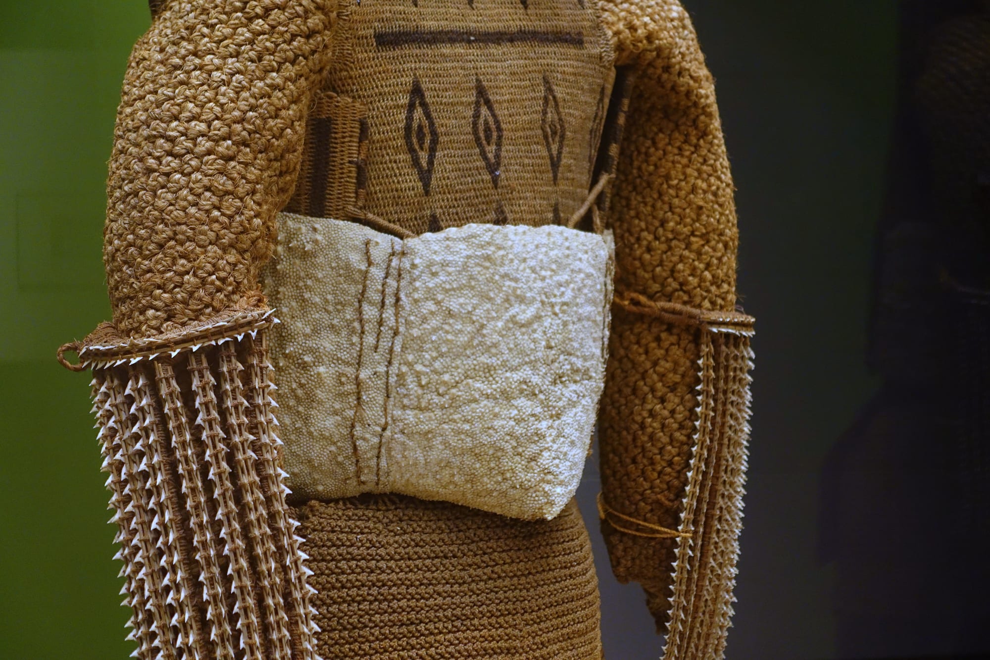 Close view of a suit of dense knotted coconut-fibre armour with dark diamond patterns on the chest, a pale textured band at the waist and spiky shark-tooth weapons hanging at each side