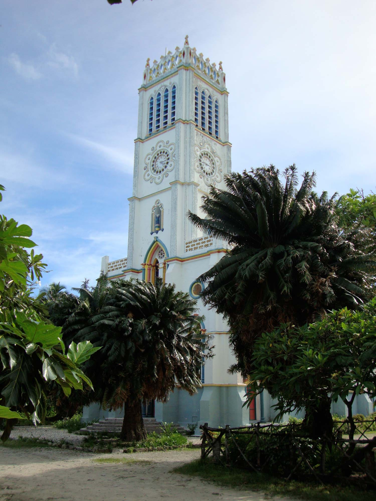 A tall white church tower with pointed windows, two ornate clock faces and a decorated crown, rising above cycad palms and a rustic wooden fence