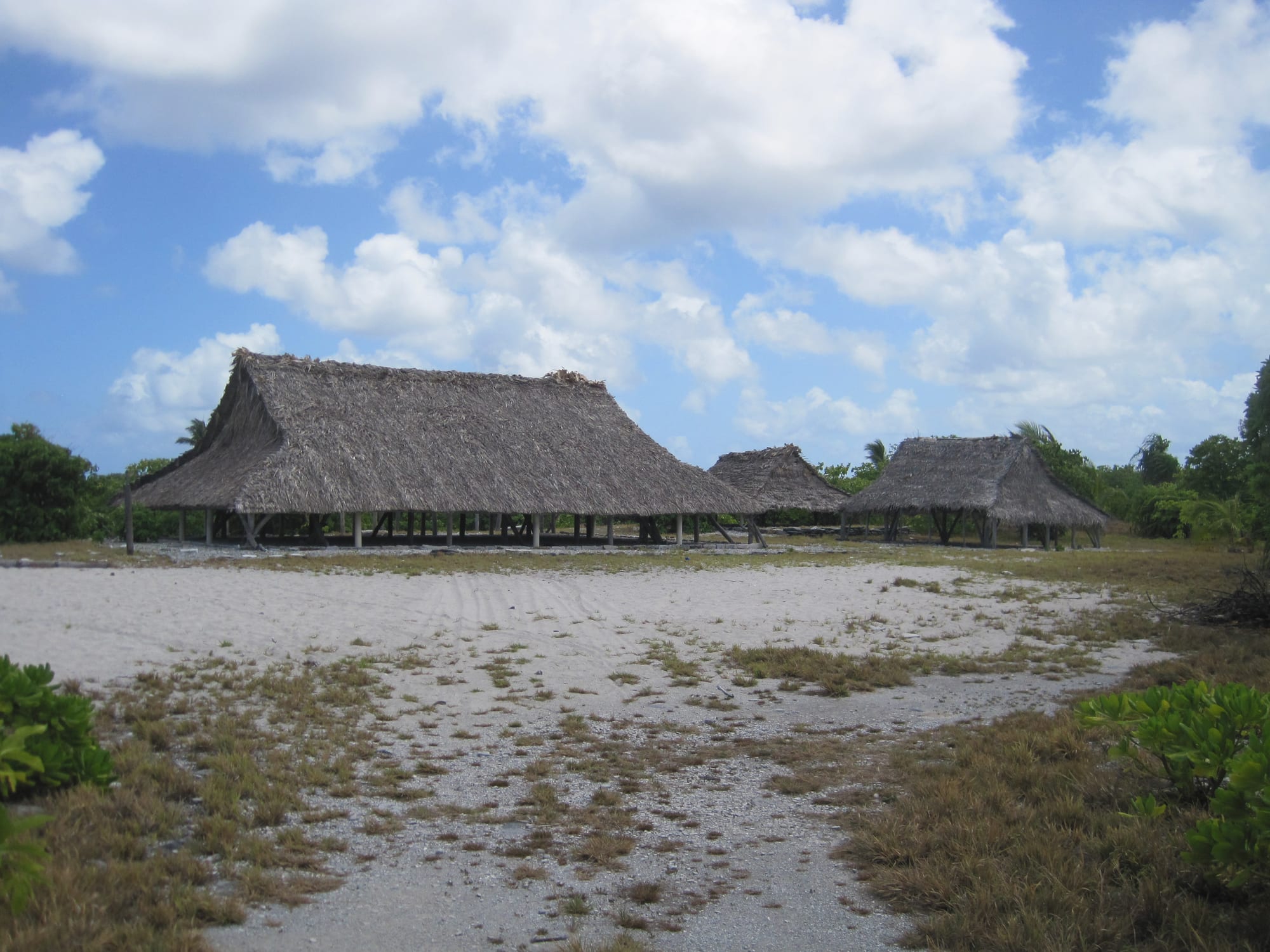 Three open-sided meeting houses with steep thatched roofs on wooden posts, set back across a wide sandy clearing under a cloudy blue sky
