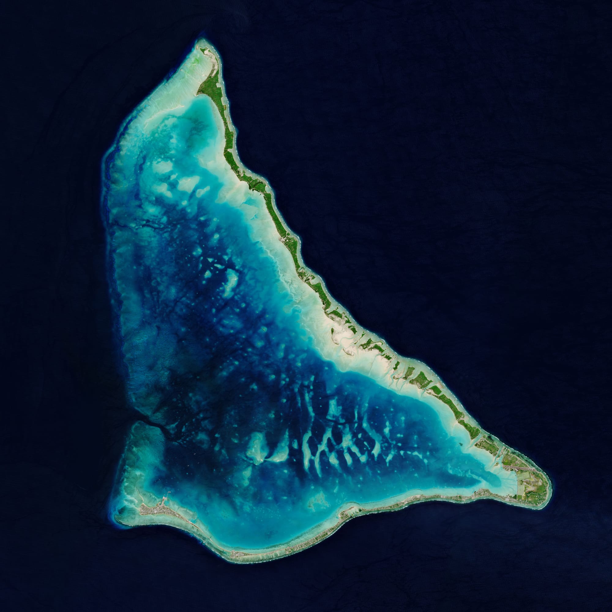 A satellite view of a narrow triangular coral atoll enclosing a lagoon in deep blue ocean