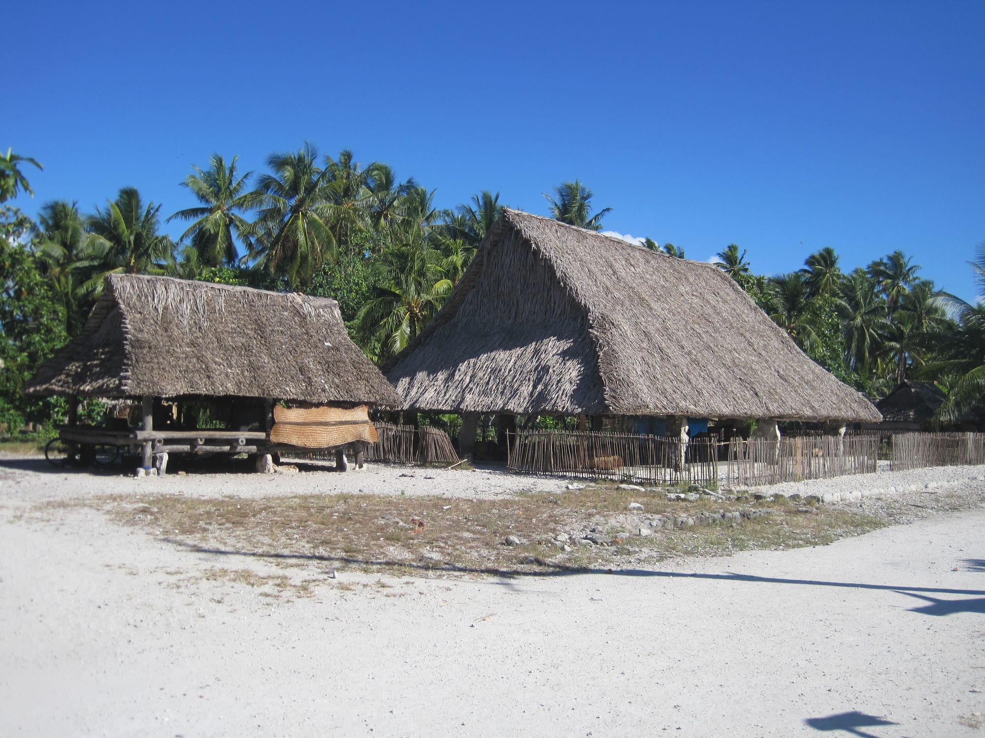 A large open-sided meeting house with a huge steep thatched roof on stone or coral posts