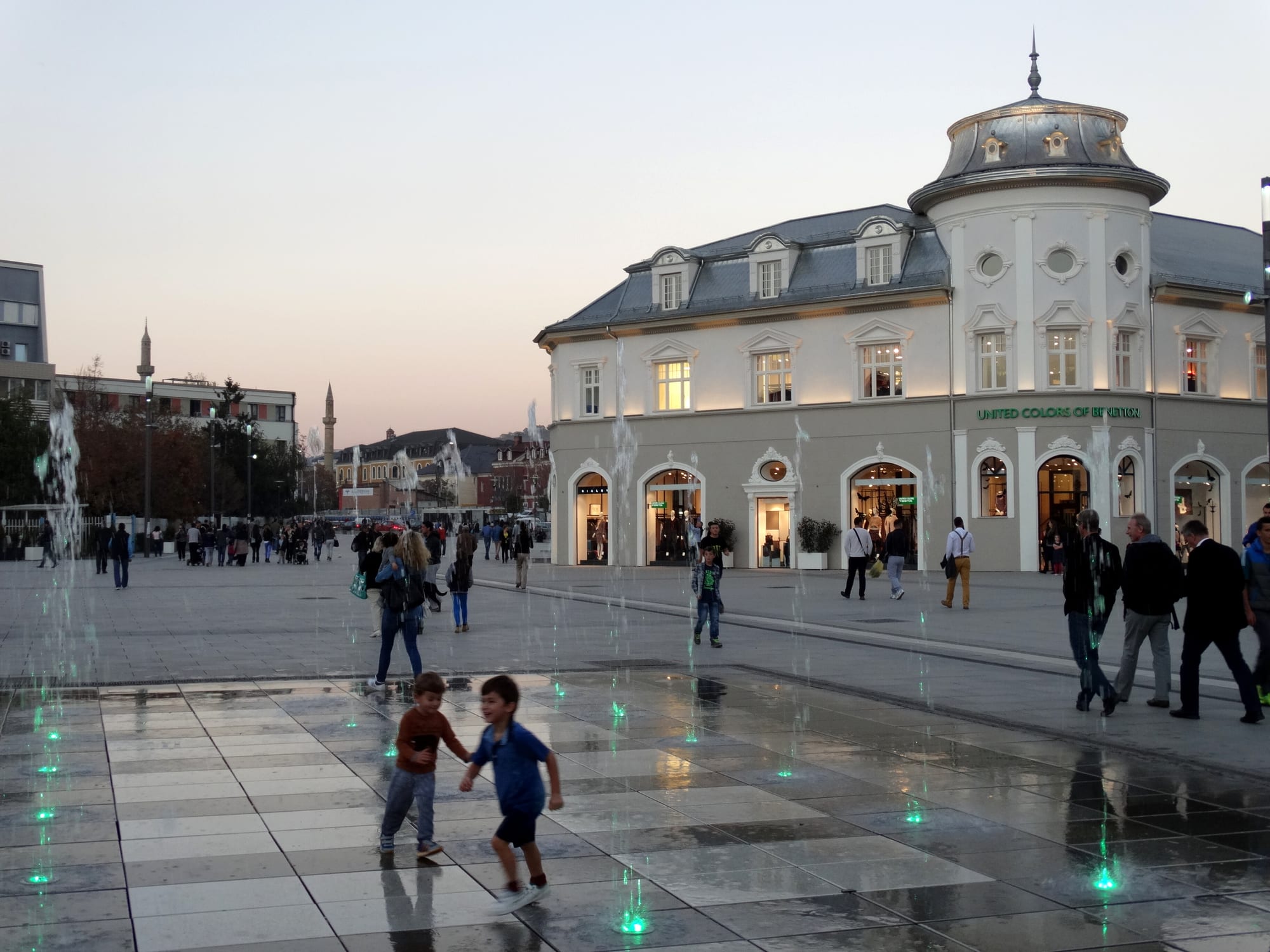 Families and children walking and playing among fountains in a paved city square at dusk, with an ornate building behind