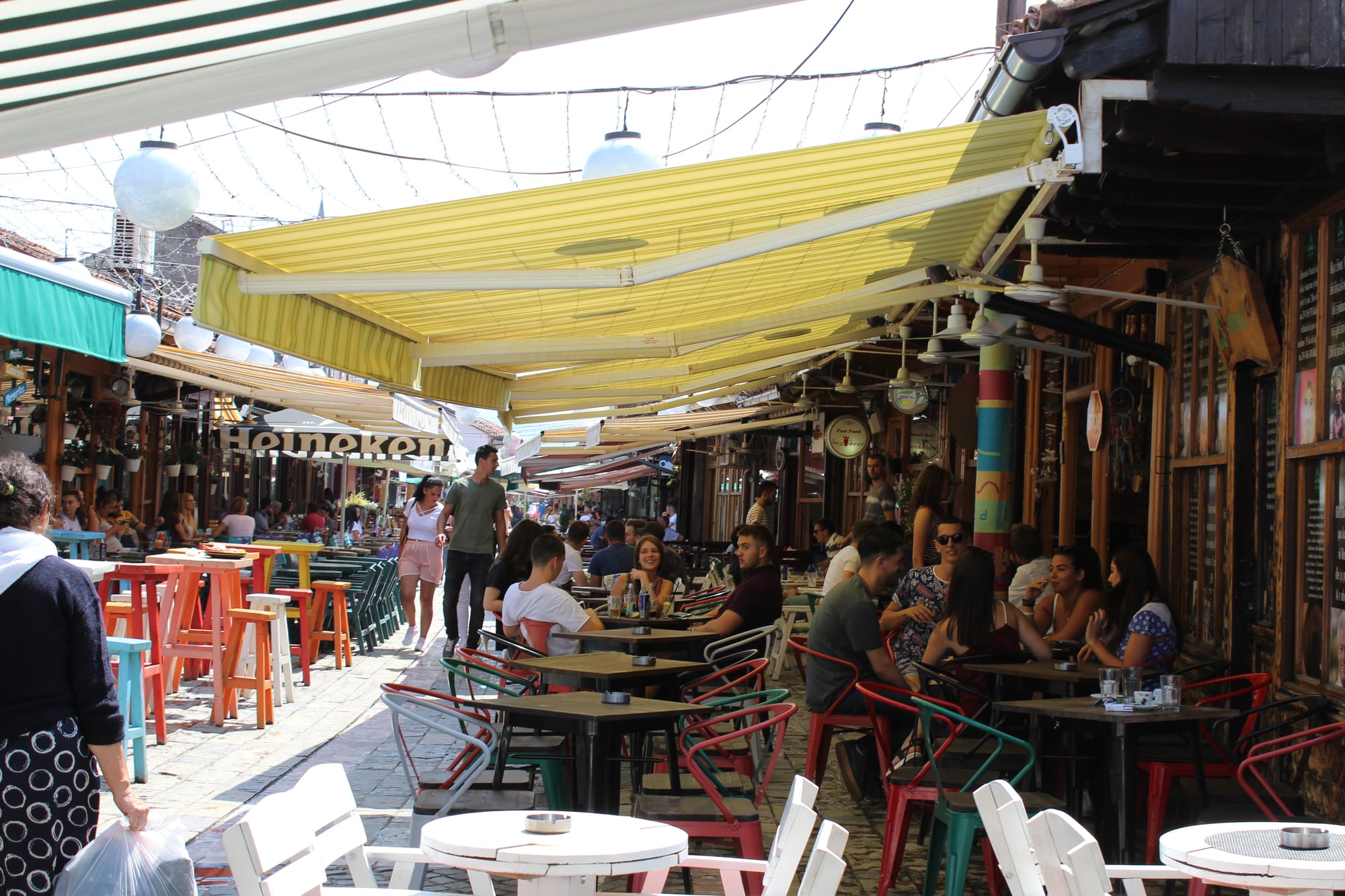 A narrow bazaar street shaded by yellow awnings, packed with people sitting at outdoor café tables and others walking through
