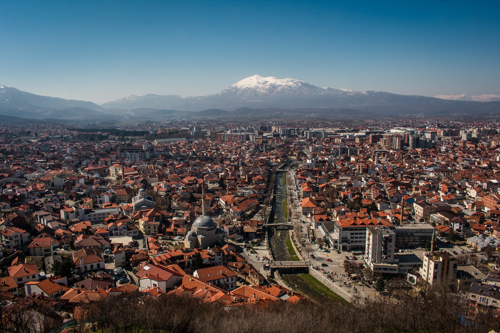 A town of red-roofed houses and minarets along a river, seen from its hilltop fortress with mountains beyond
