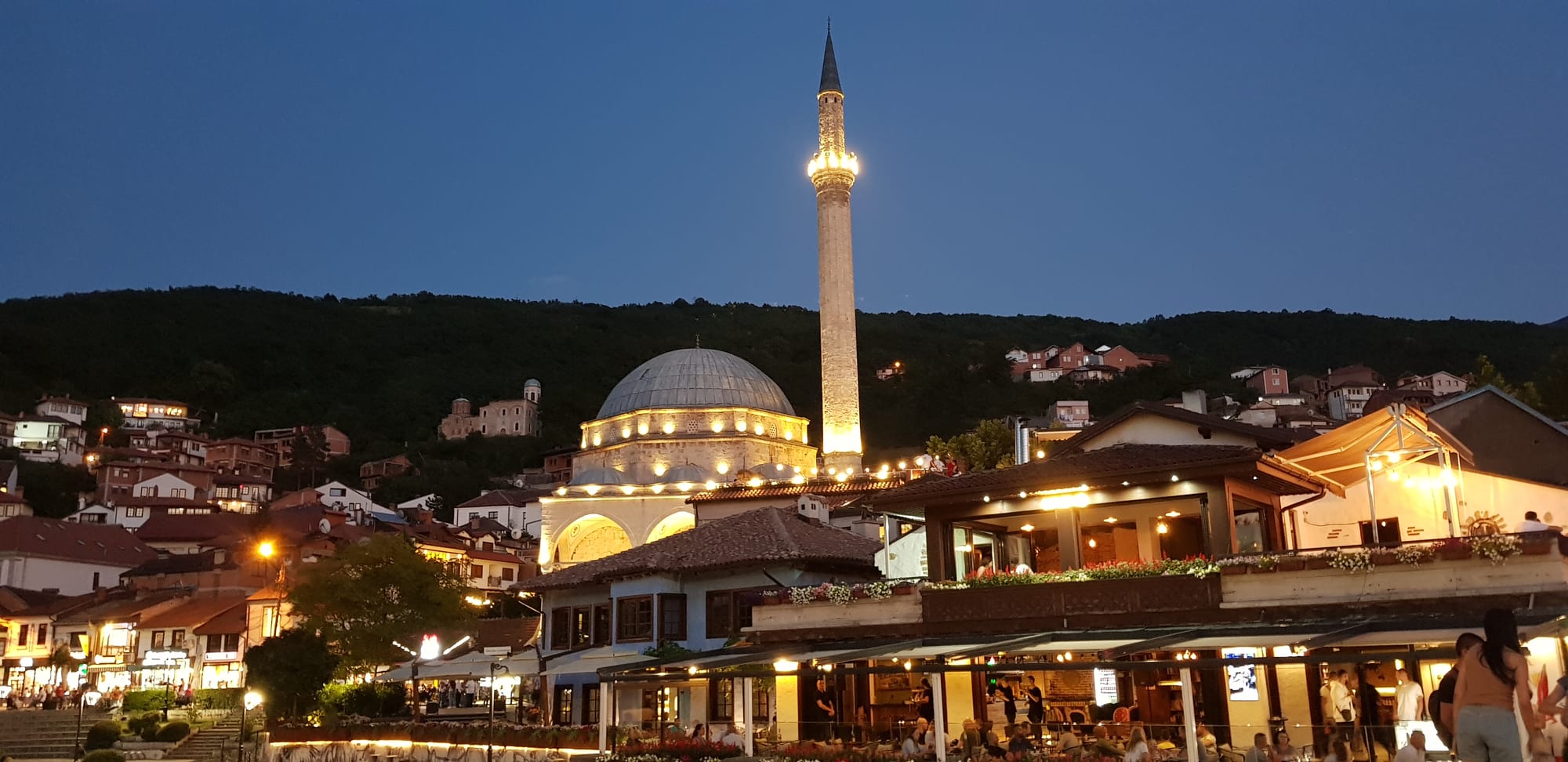 A domed mosque with a tall lit minaret rising above rooftops and busy riverside café terraces at dusk, with a wooded hillside behind