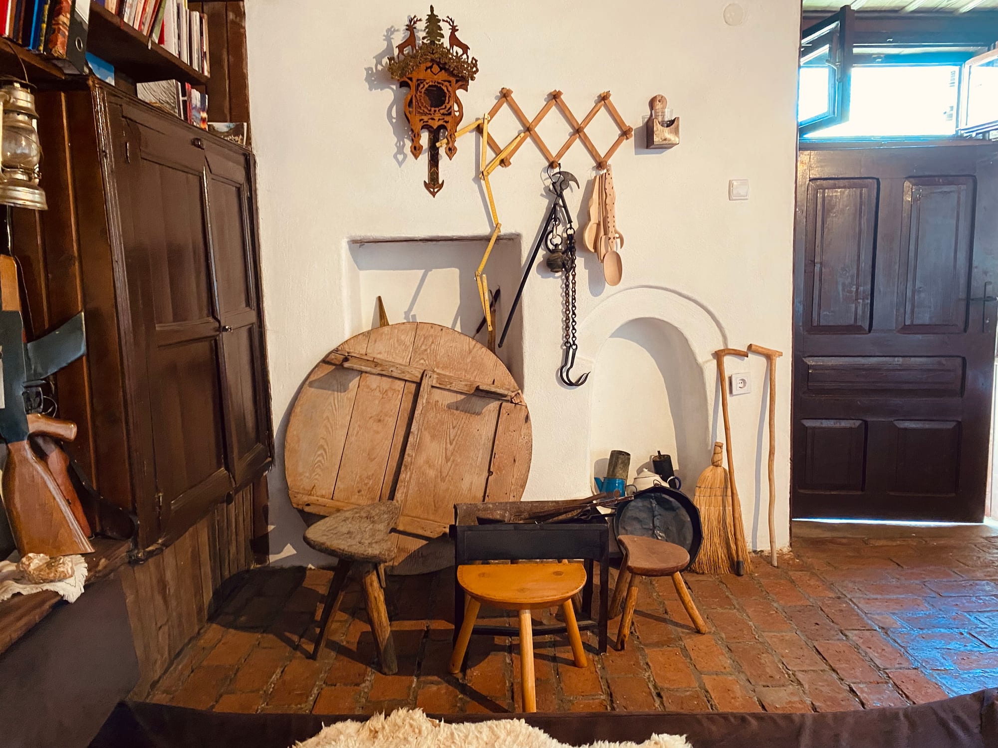 A whitewashed room with a brick floor, low wooden stools, a folding round wooden table, a broom, walking sticks and iron hooks hanging on the wall beside a panelled wooden door