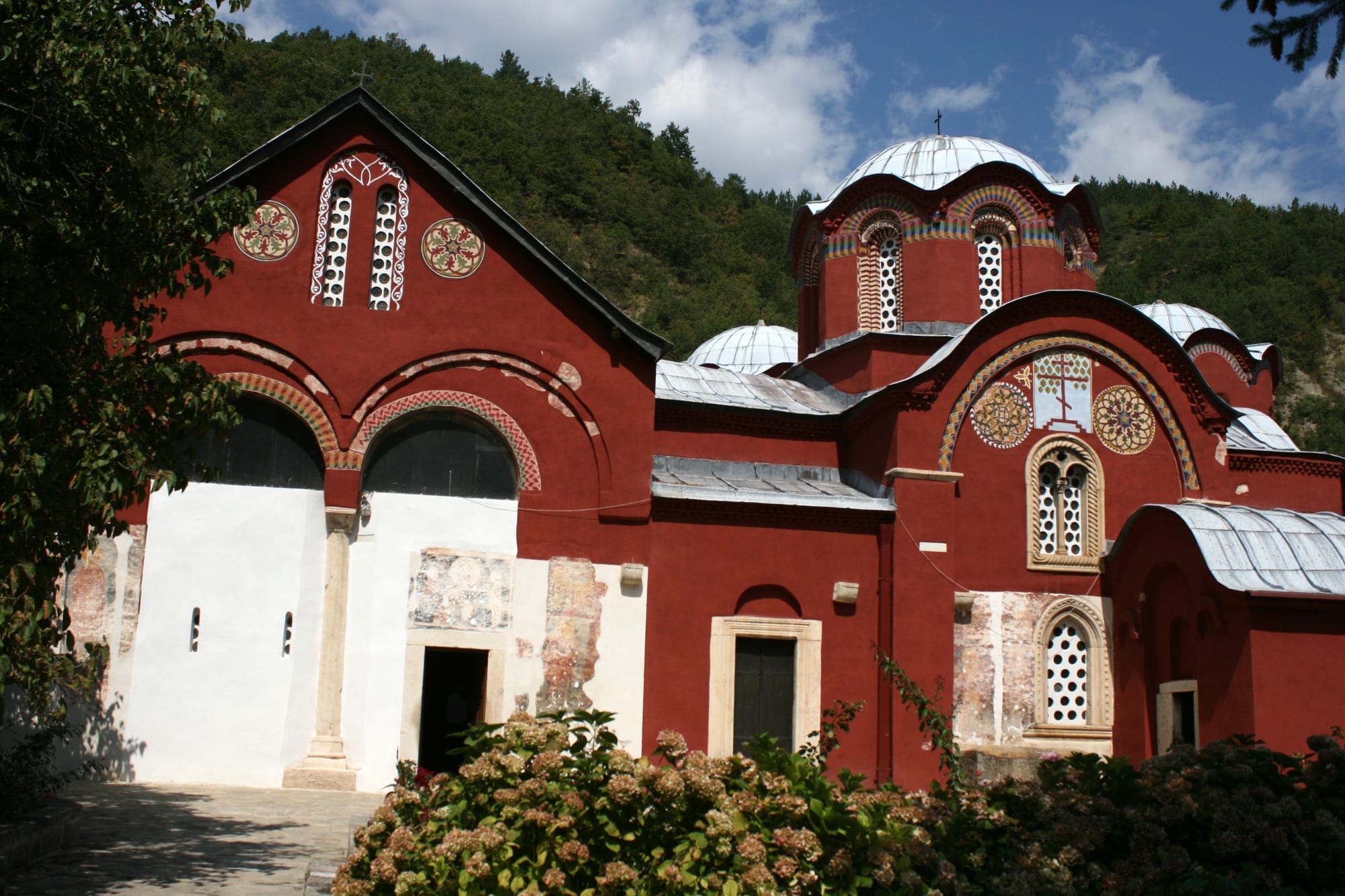 A red-painted Orthodox church complex with grey domes and patterned round windows, set below a wooded hillside
