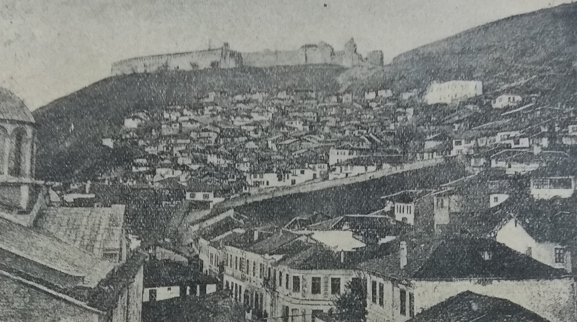 An old town of red roofs and minarets along a river below a hilltop fortress