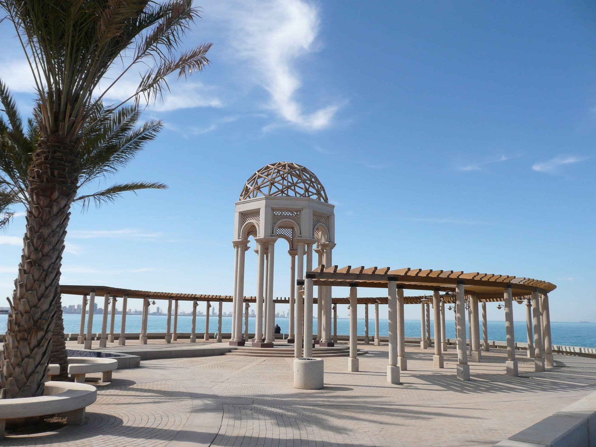 A domed stone pavilion and covered walkway along the Salmiya waterfront promenade, with the Kuwait City skyline visible across the bay