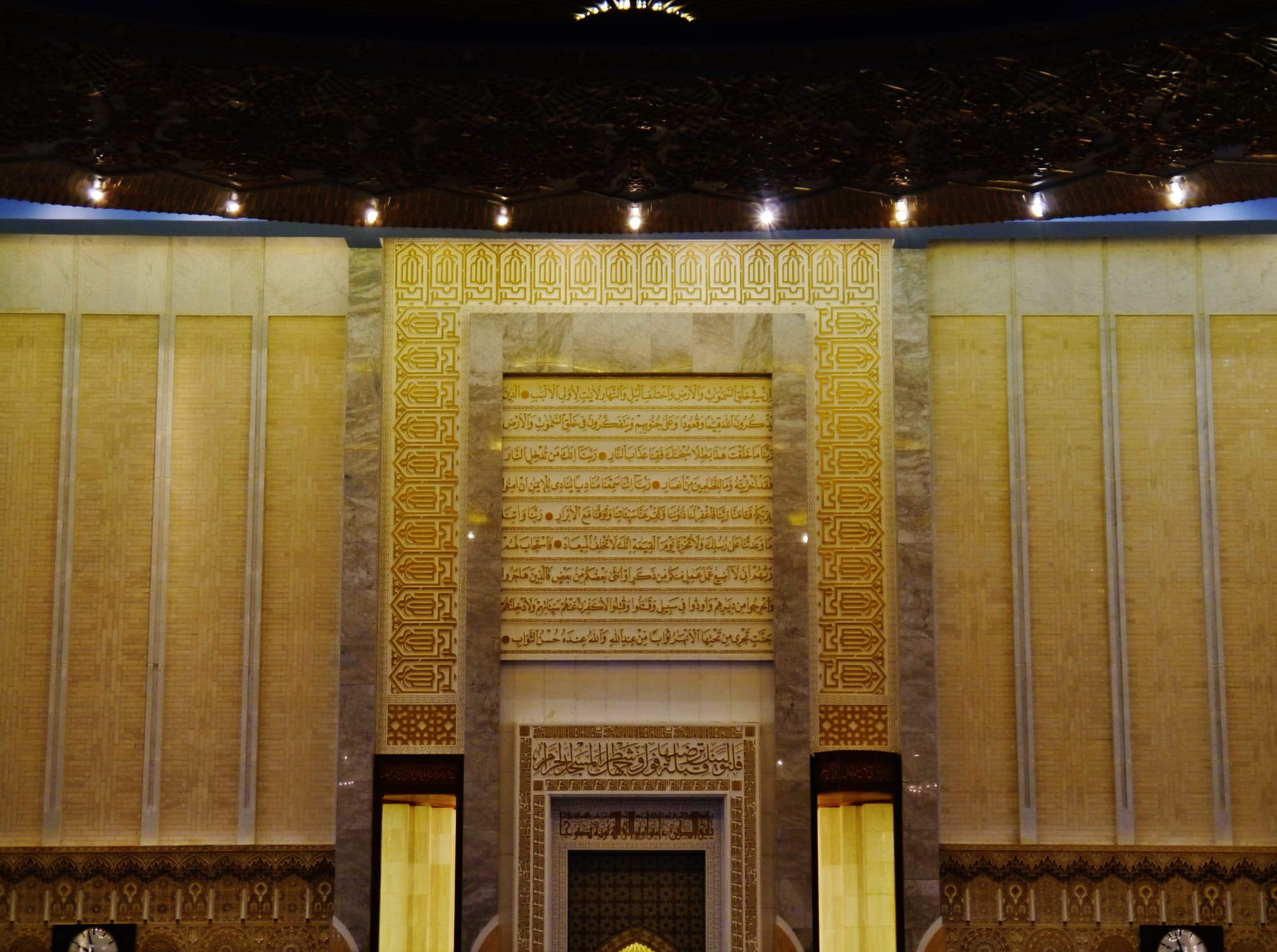A gold-inscribed mihrab niche and marble wall inside Kuwait's Grand Mosque, lit by rows of small ceiling lights