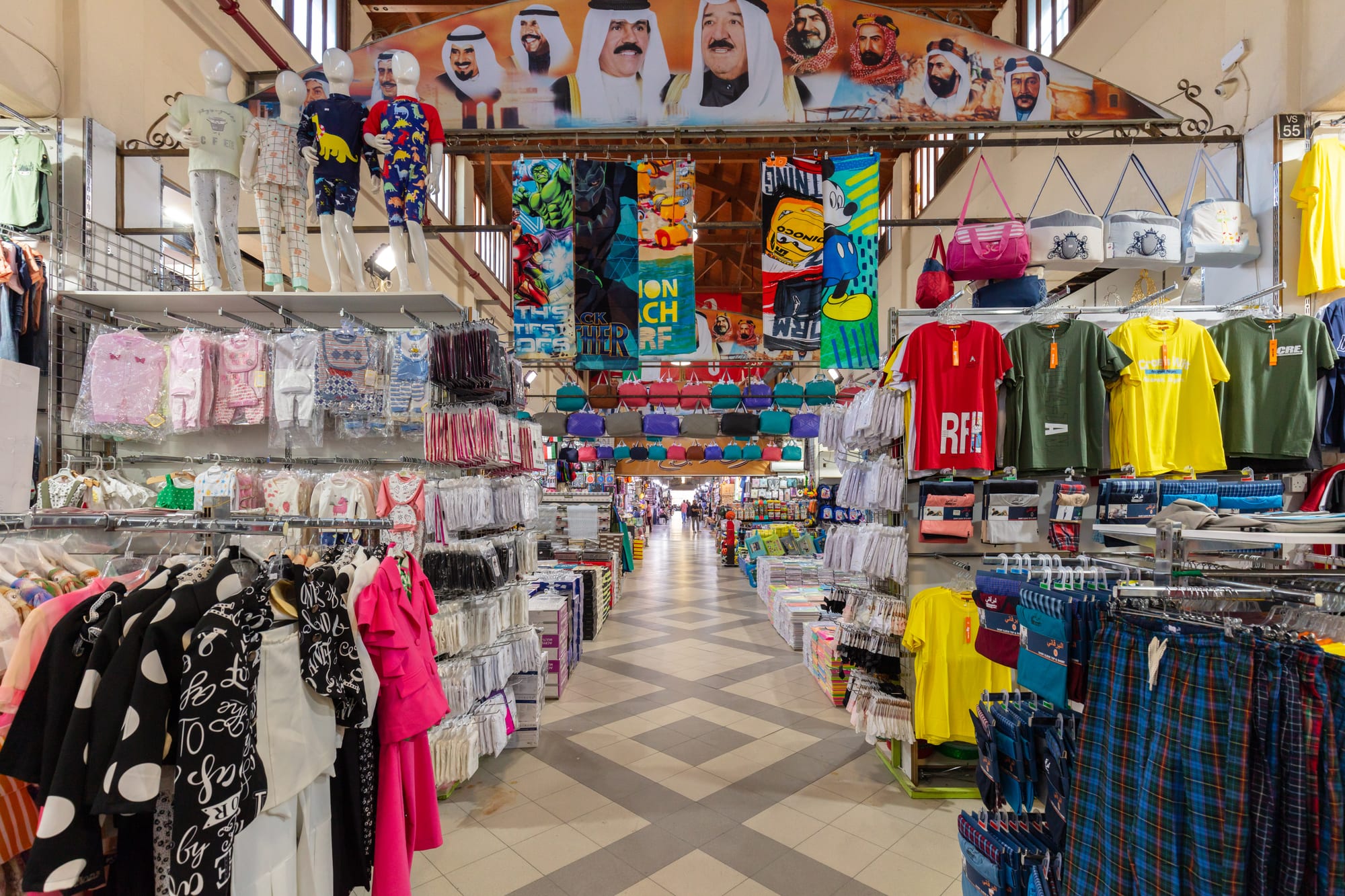 A covered market aisle lined with clothing stalls and hanging goods, portraits of Kuwait's rulers displayed above the entrance
