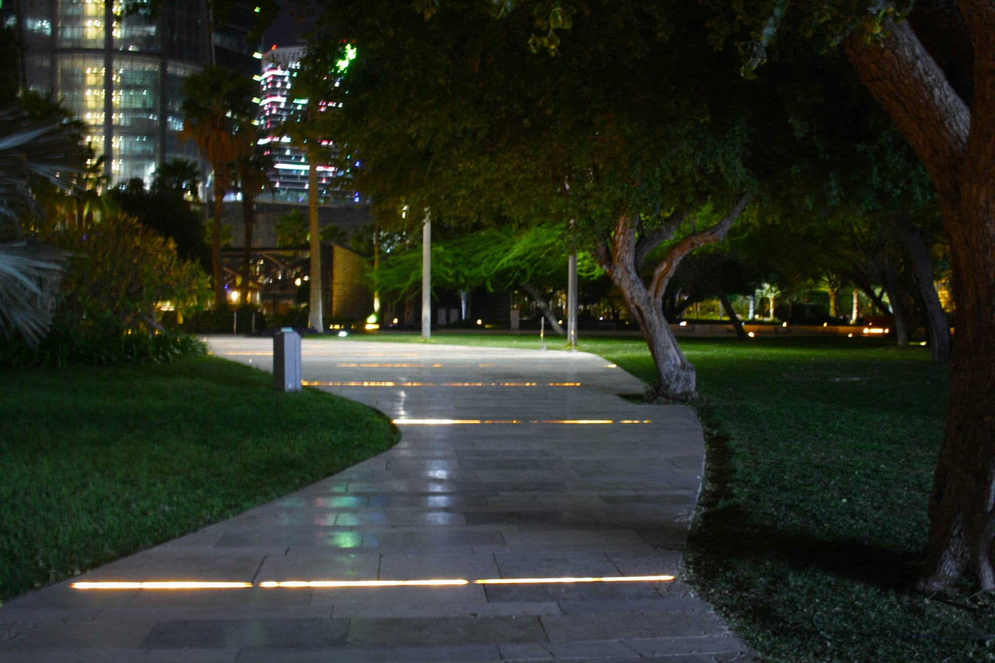 A landscaped park path lined with trees and lights, city towers behind