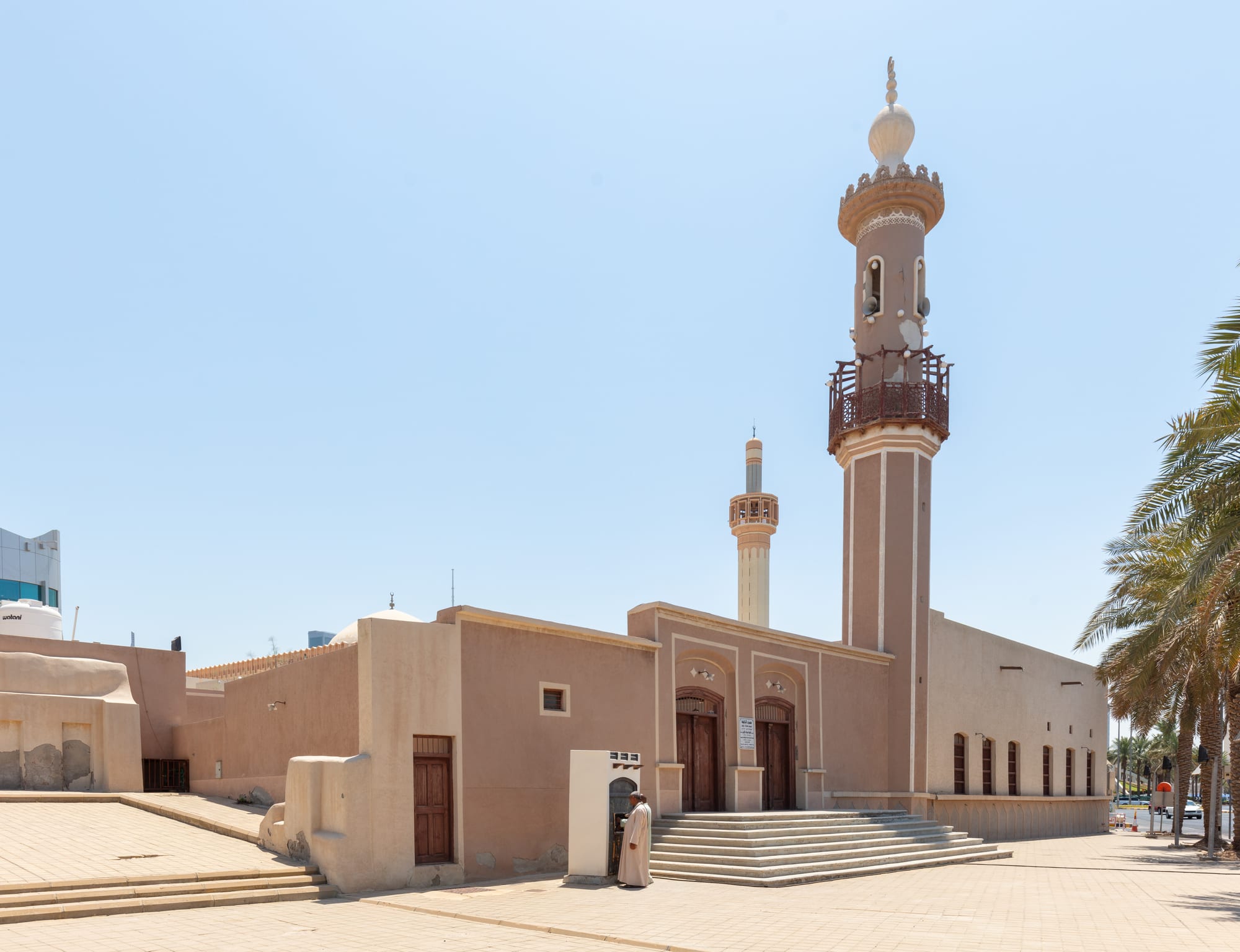 A low sand-coloured mosque with wooden double doors at the top of a flight of steps, a tall minaret with a carved wooden balcony, a second slimmer minaret behind, and palm trees on the right under a pale blue sky