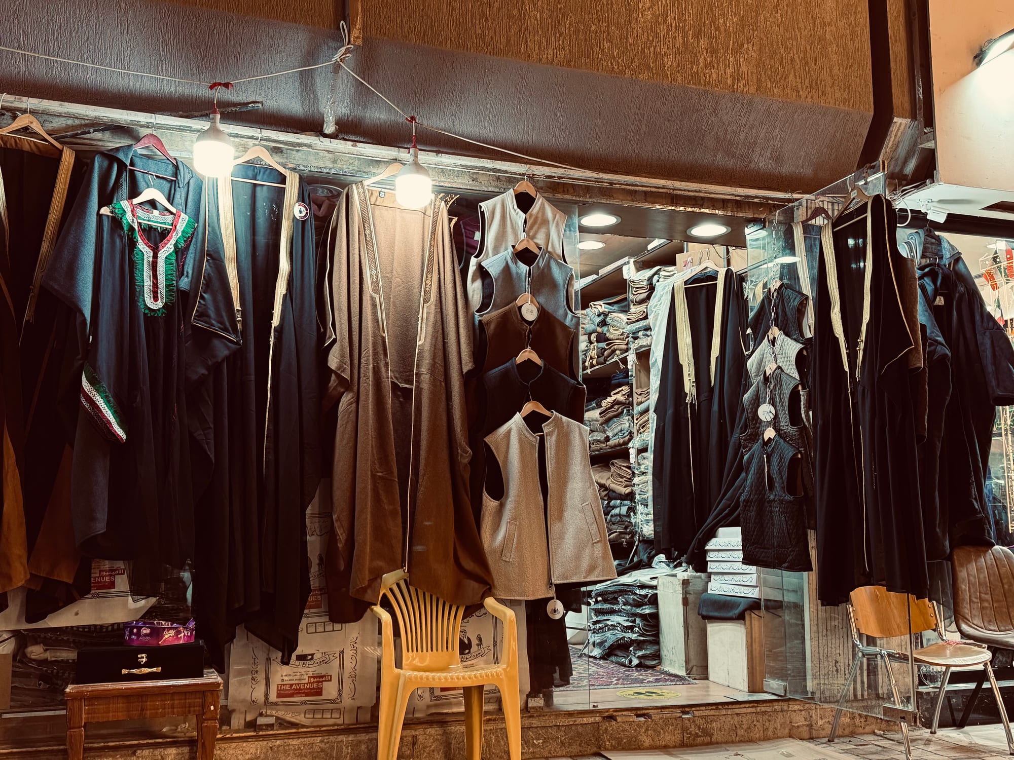 A shopfront hung with black, brown and cream cloaks edged with gold braid, alongside waistcoats on hangers, lit by bare bulbs, with a plastic chair in front