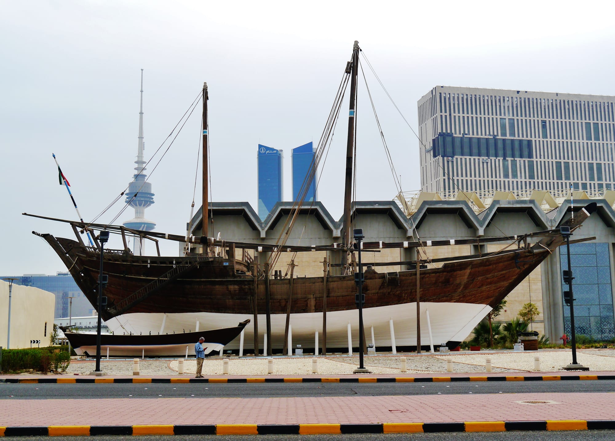 A large wooden dhow with two tall masts displayed on a white stand beside a road, with a smaller boat in front and modern towers and a telecommunications tower behind
