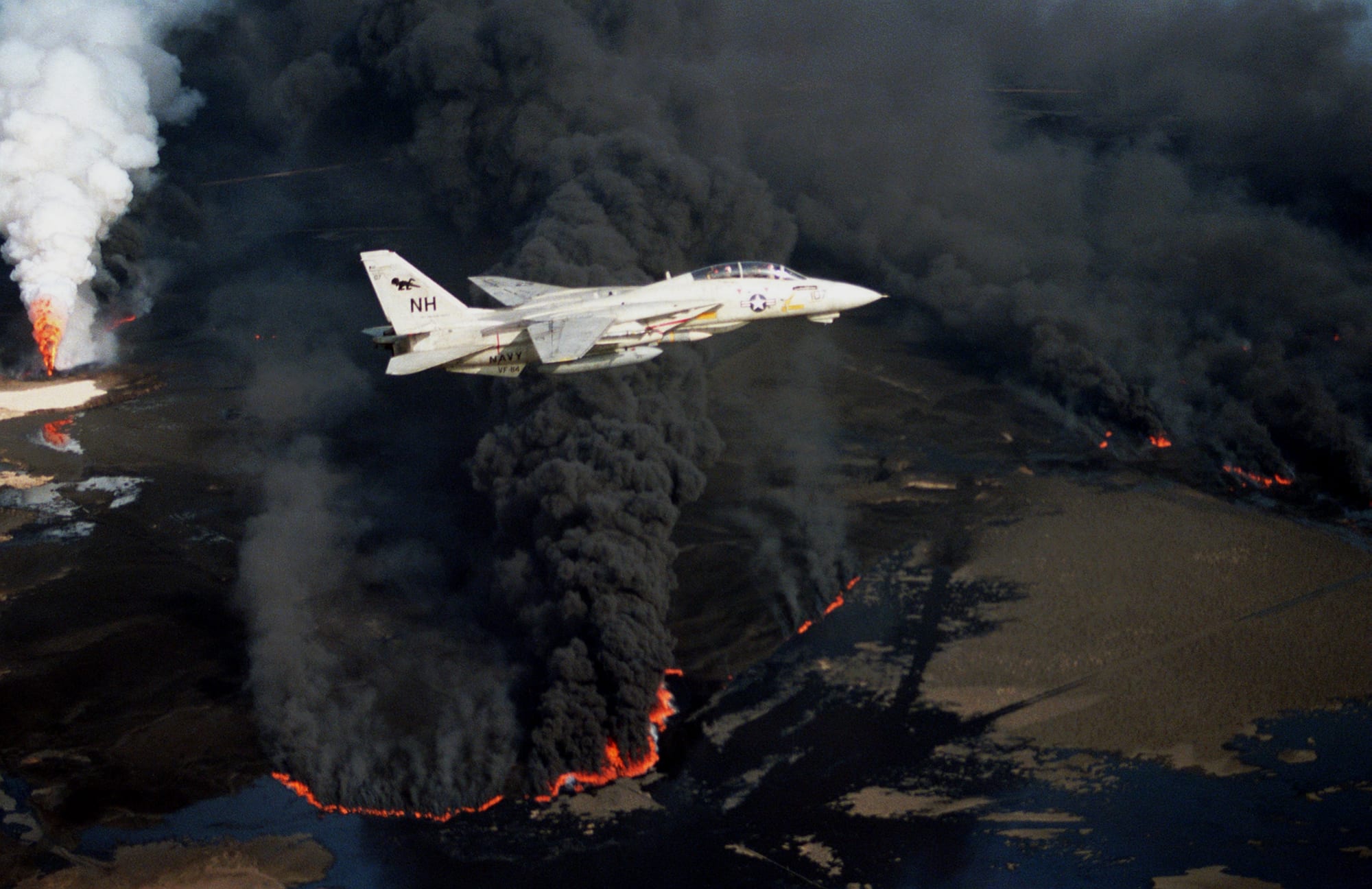 A US Navy F-14 fighter flying over black smoke from a burning Kuwaiti oil well, 1991