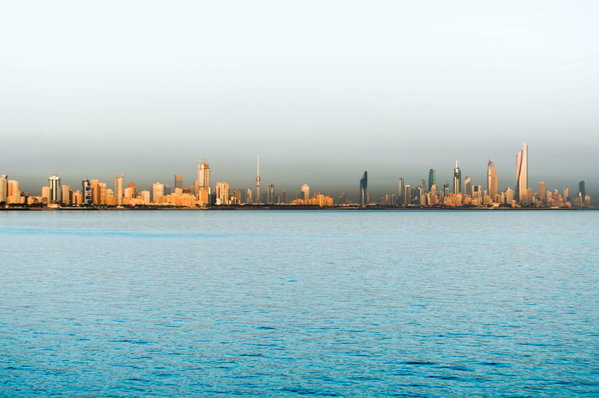 A waterfront skyline of glass towers and the pointed Kuwait Towers along a blue Gulf coast
