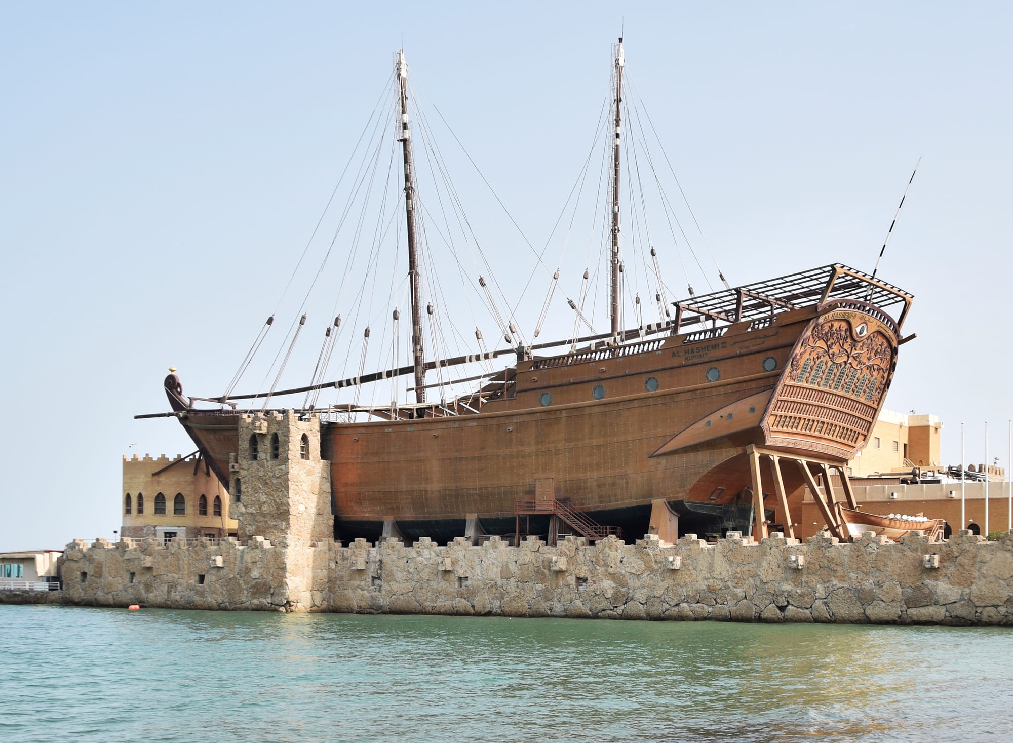 A very large wooden dhow with two masts and a carved stern, raised on supports behind a low stone sea wall at the water's edge