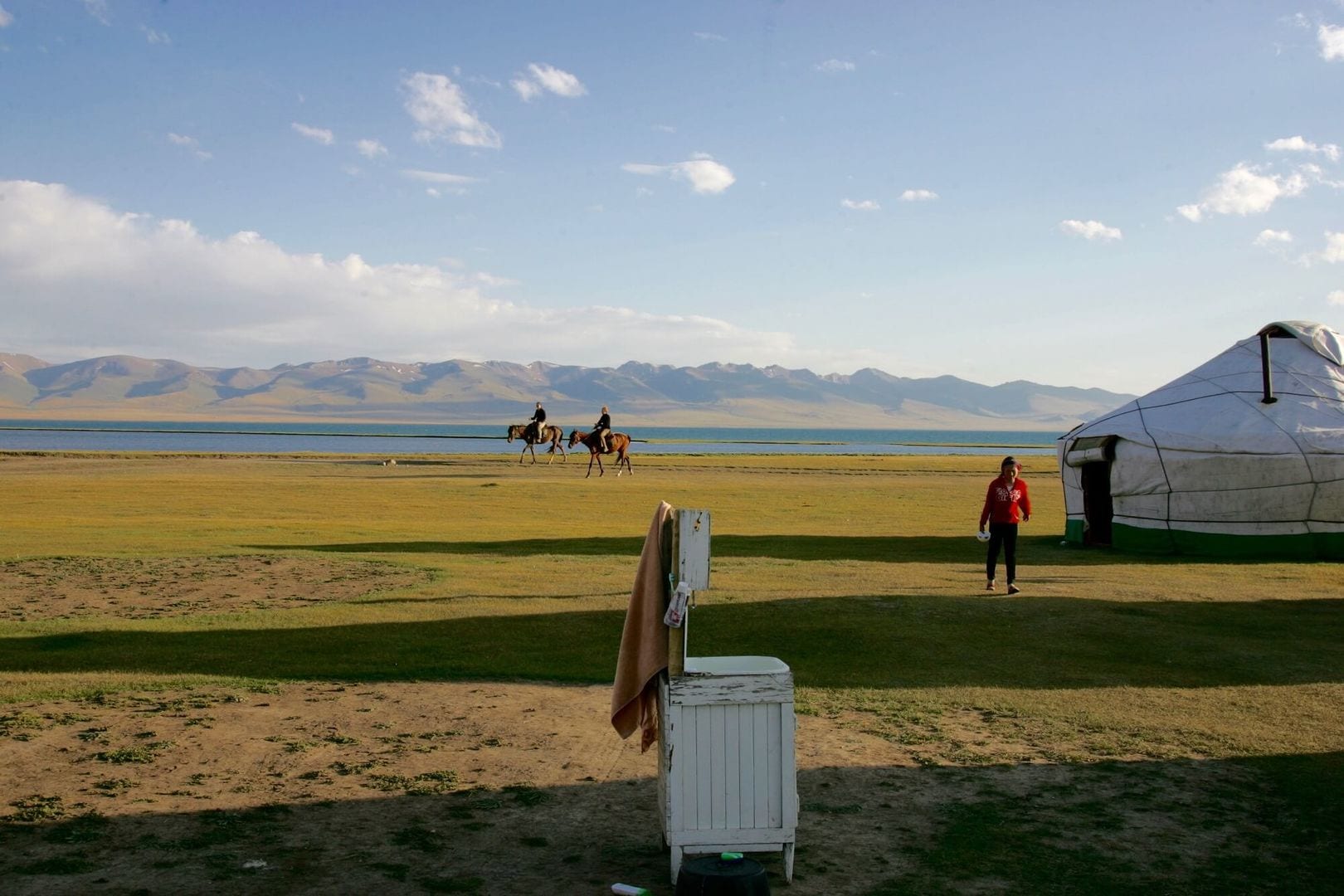 Horses grazing beside yurts on open grassland by a lake, mountains behind