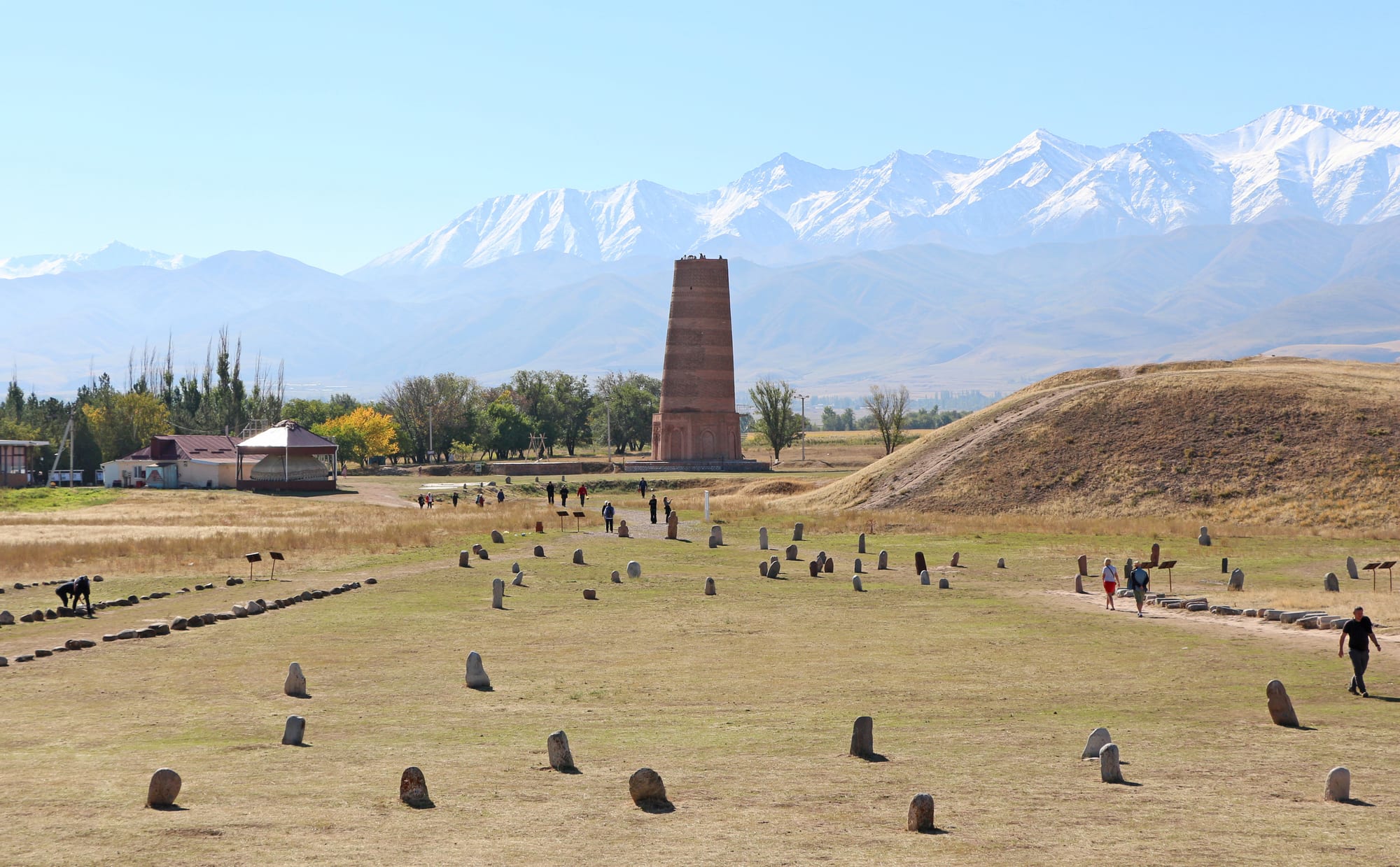 A stout, tapering brick minaret standing alone on a grassy plain with mountains behind