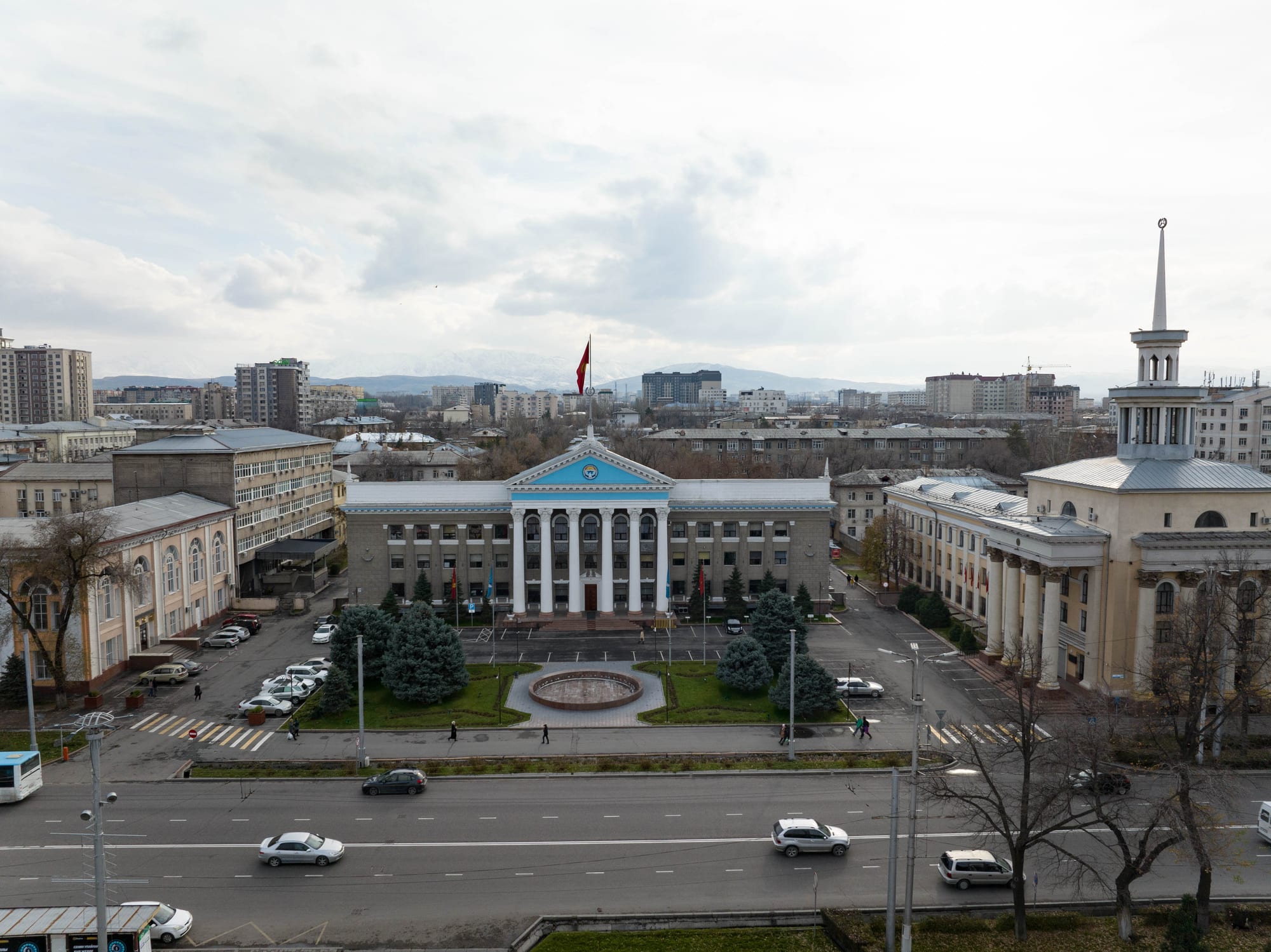An aerial view of Bishkek's columned city hall and surrounding streets, snow-capped mountains behind