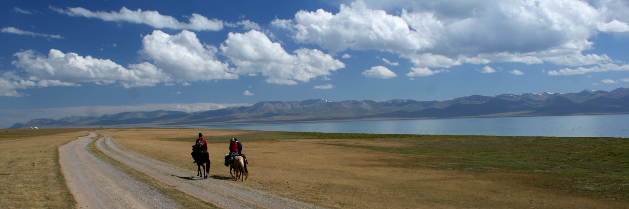 Two horseback riders on a track beside a wide alpine lake under a big sky