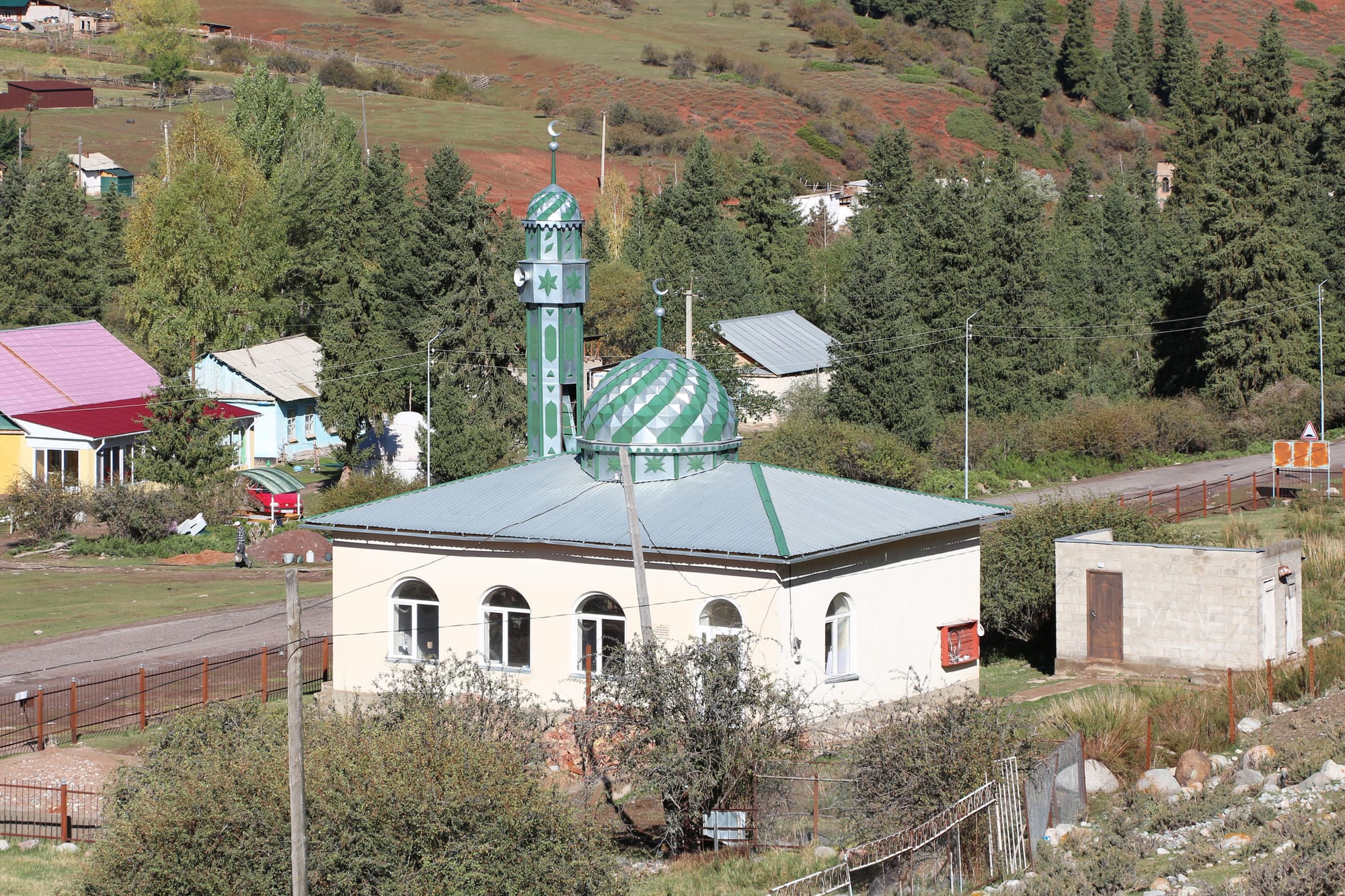 A small white mosque with a green-striped dome and a slender green minaret topped by a crescent, among wooden houses and tall fir trees on a hillside