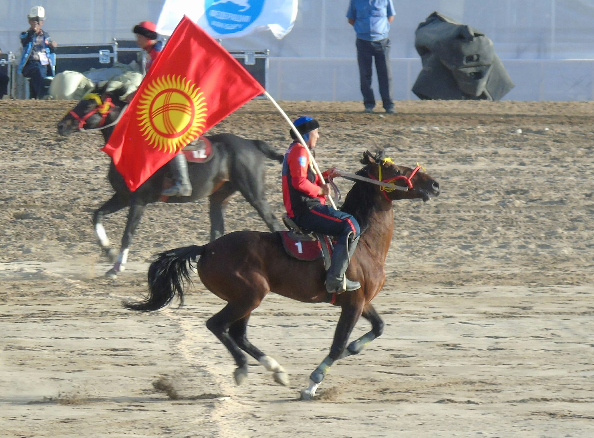 A rider in a red and blue jersey gallops a brown horse across a dusty field holding a large red Kyrgyz flag, with a second horseman behind him