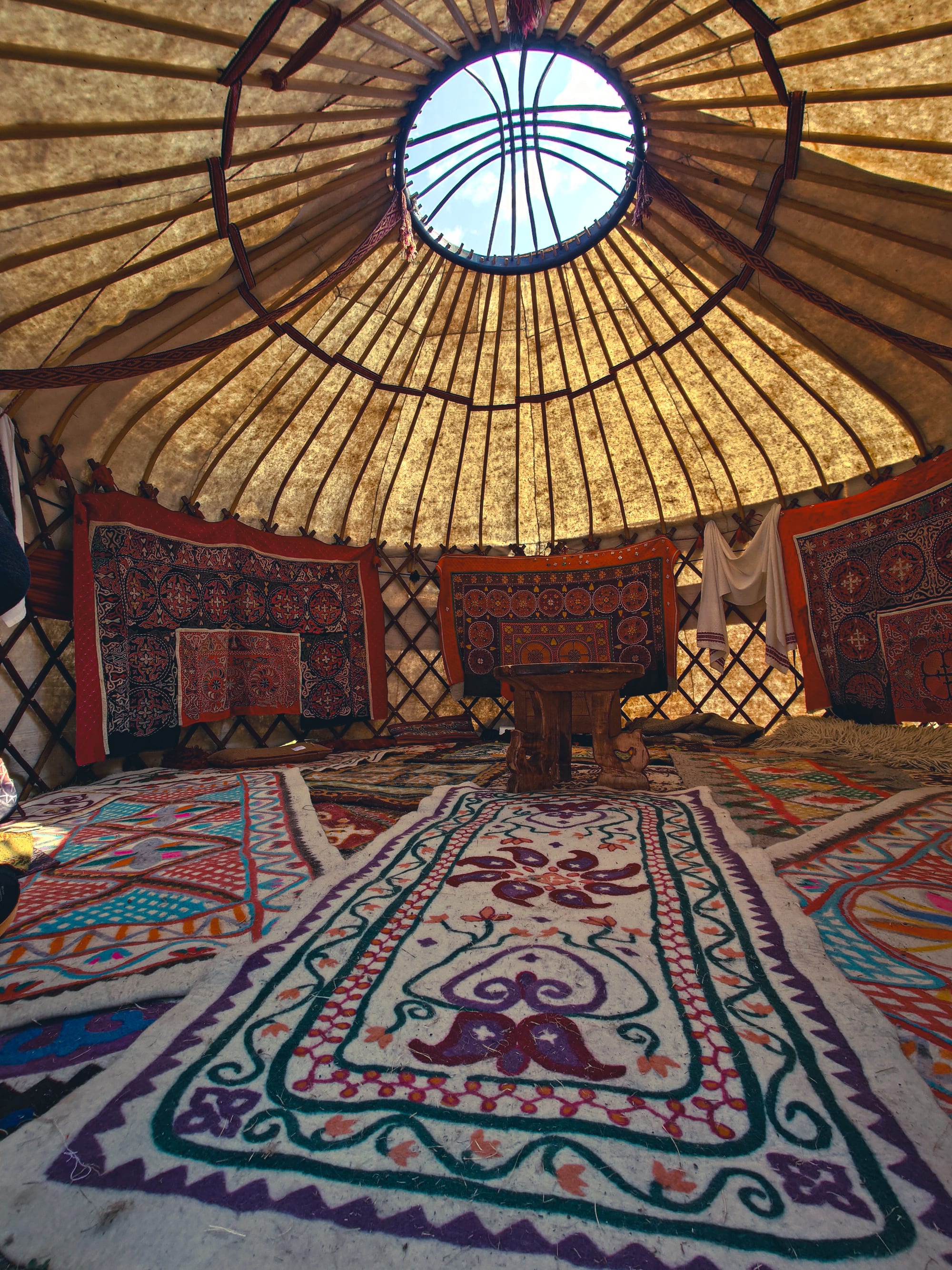 The interior of a yurt seen from floor level, with colourful felt carpets covering the floor and walls, a low wooden table, and the roof poles converging on a round opening to the sky