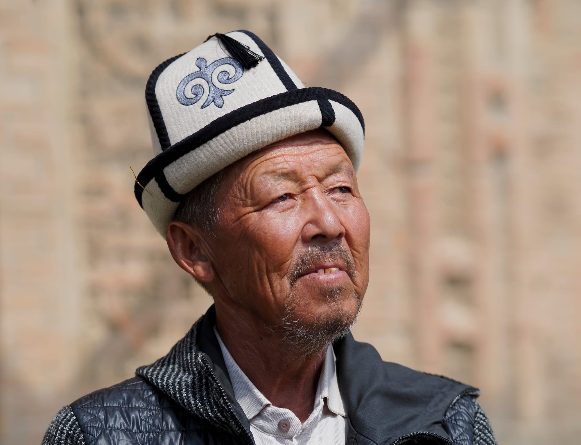 An older man with a grey beard wearing a tall white felt hat with black trim, an embroidered blue motif and a black tassel on top