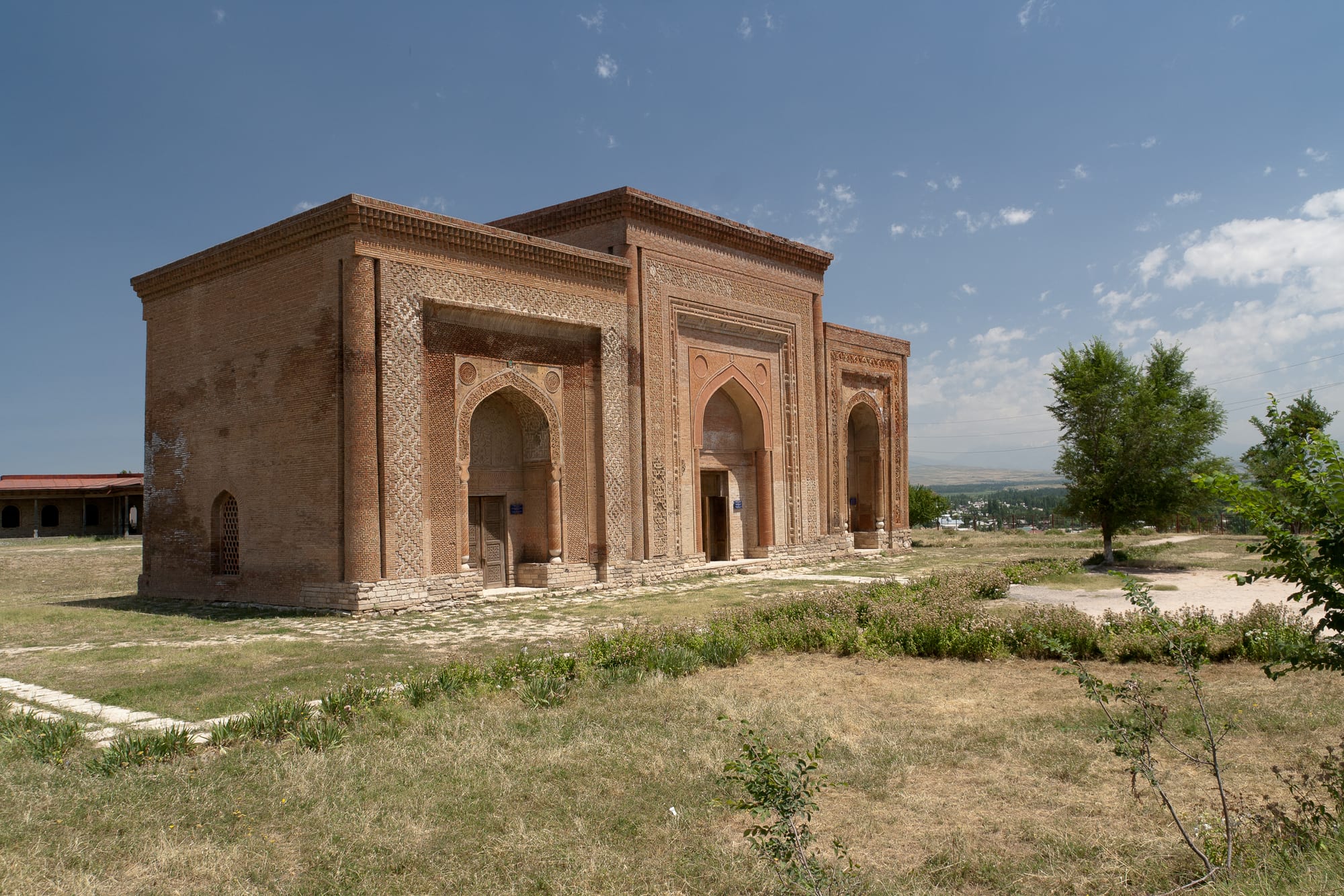 A long brick mausoleum with three pointed-arch portals framed by bands of carved geometric brickwork, standing on dry grass under a blue sky