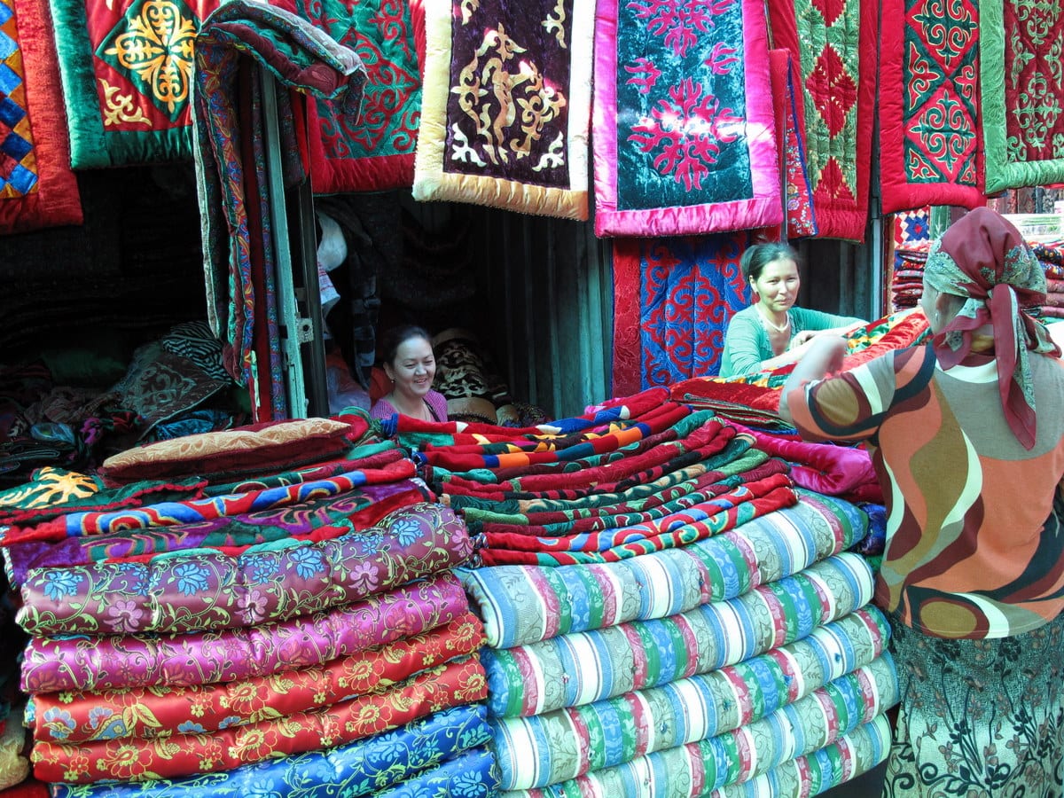 Stalls piled with brightly patterned quilted blankets and cushions at a covered market, two vendors seated behind the display talking with a customer