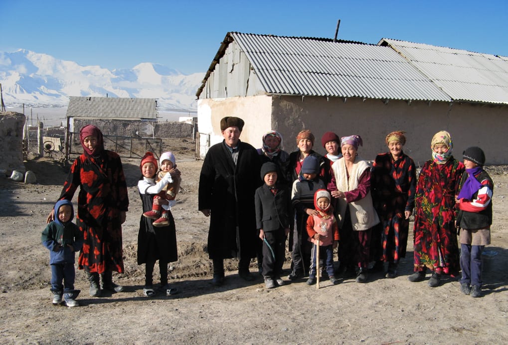A large extended Kyrgyz family, from young children to grandparents, standing together outside a house with snow-capped mountains behind