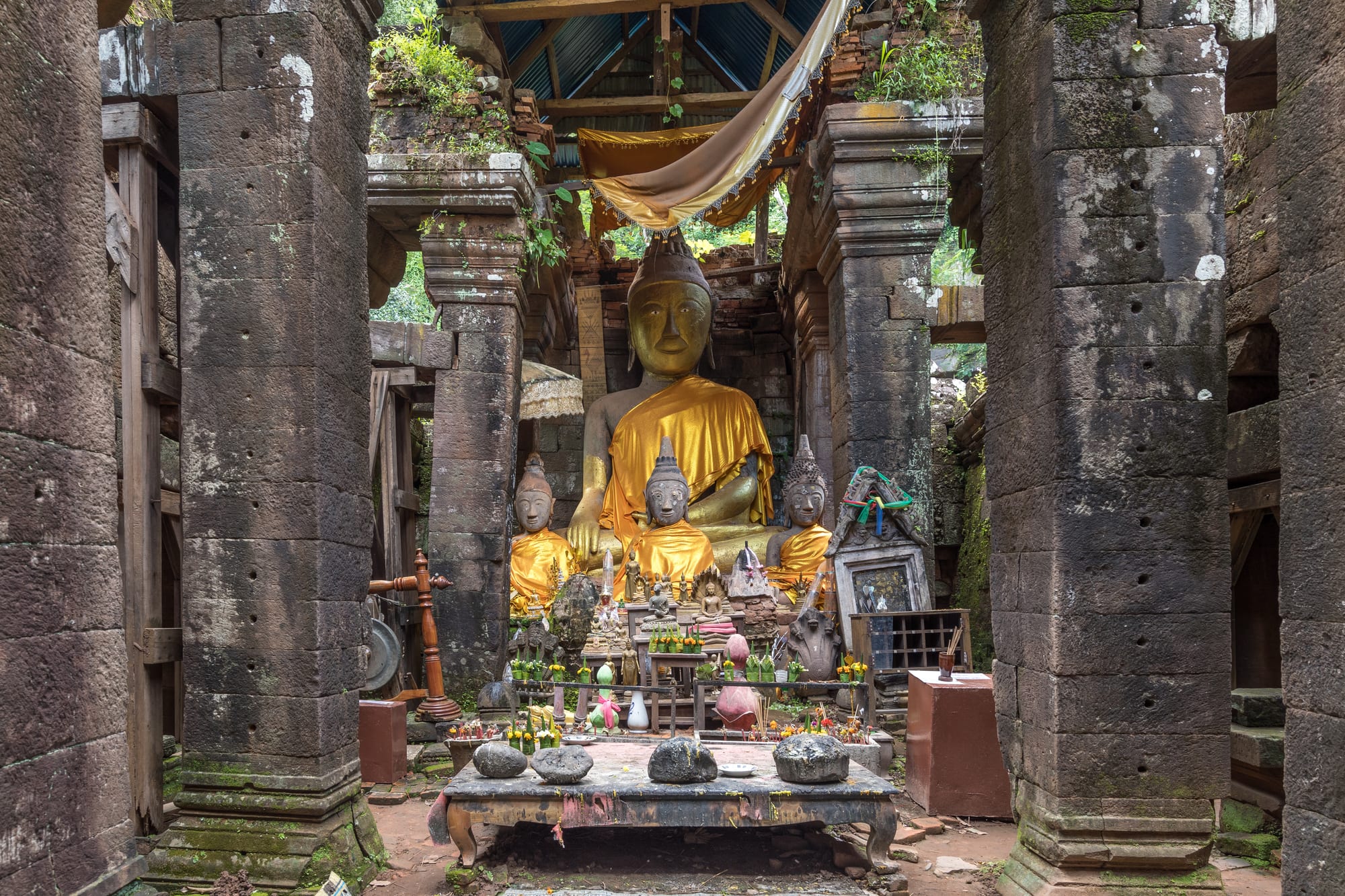 Ruined stone temple terraces climbing a jungle-covered hillside, statues draped in orange cloth