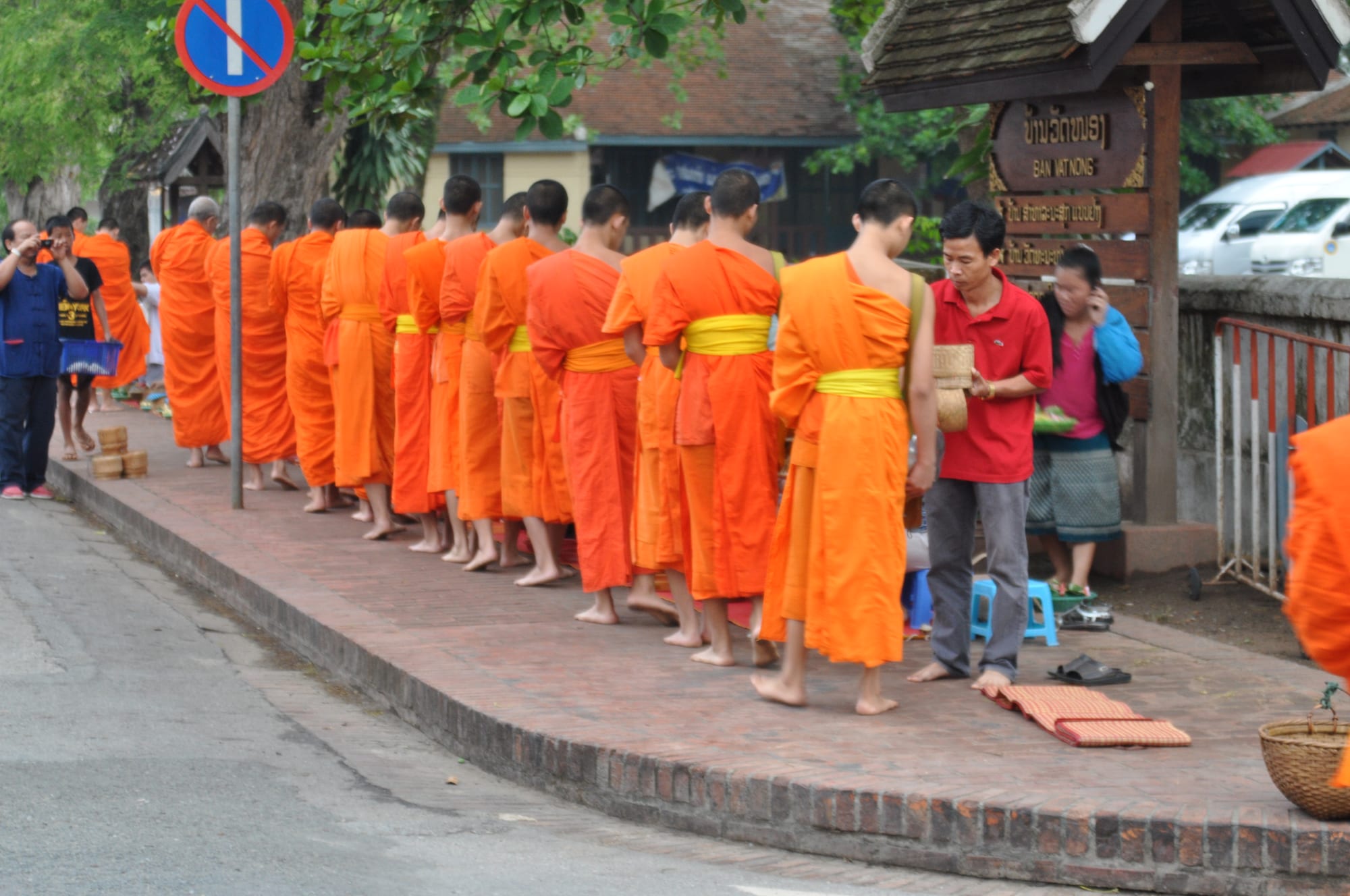 A line of saffron-robed monks walking with alms bowls past kneeling townspeople at dawn