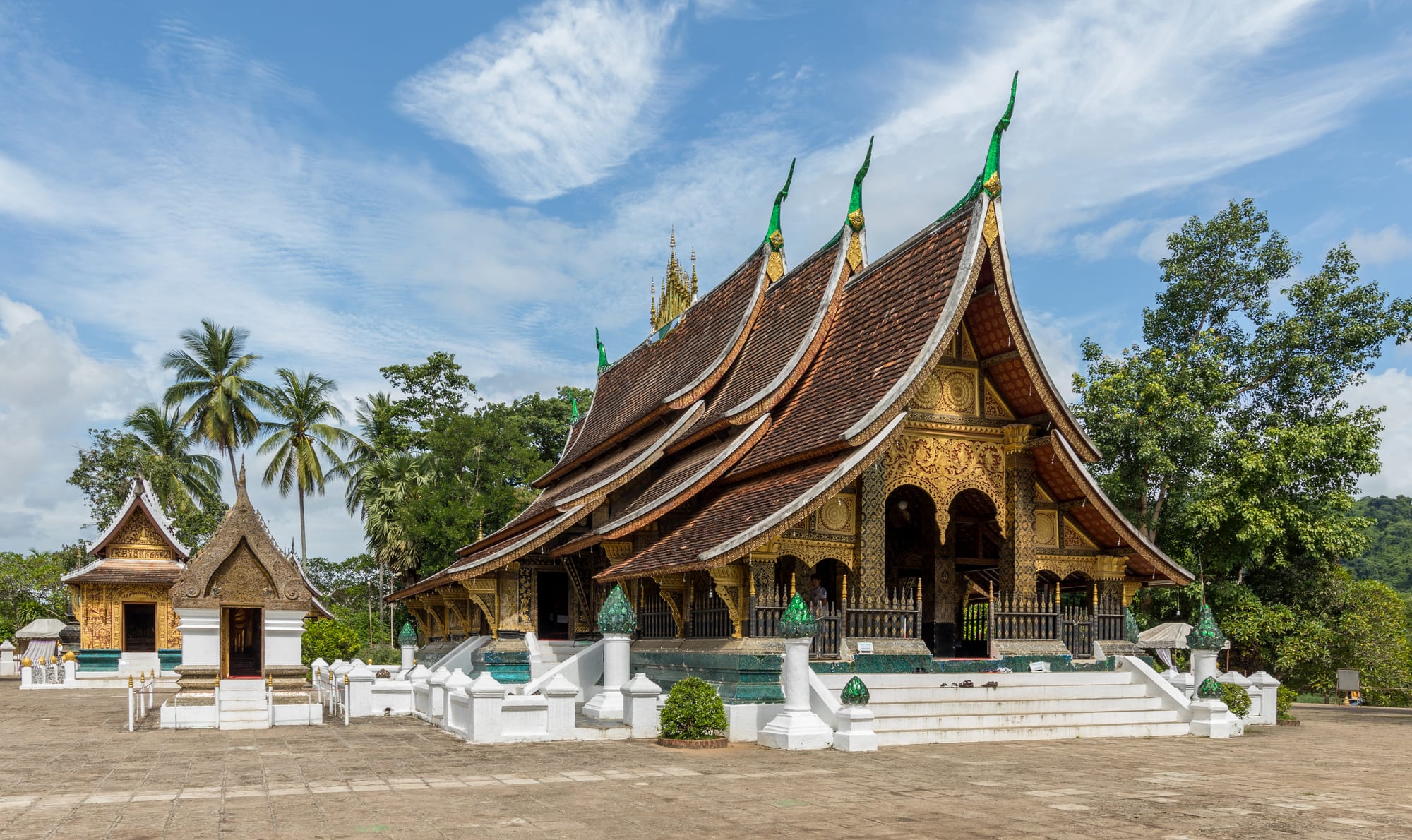 An ornate low temple with a steep tiered roof and gold-stencilled walls, palm trees around it