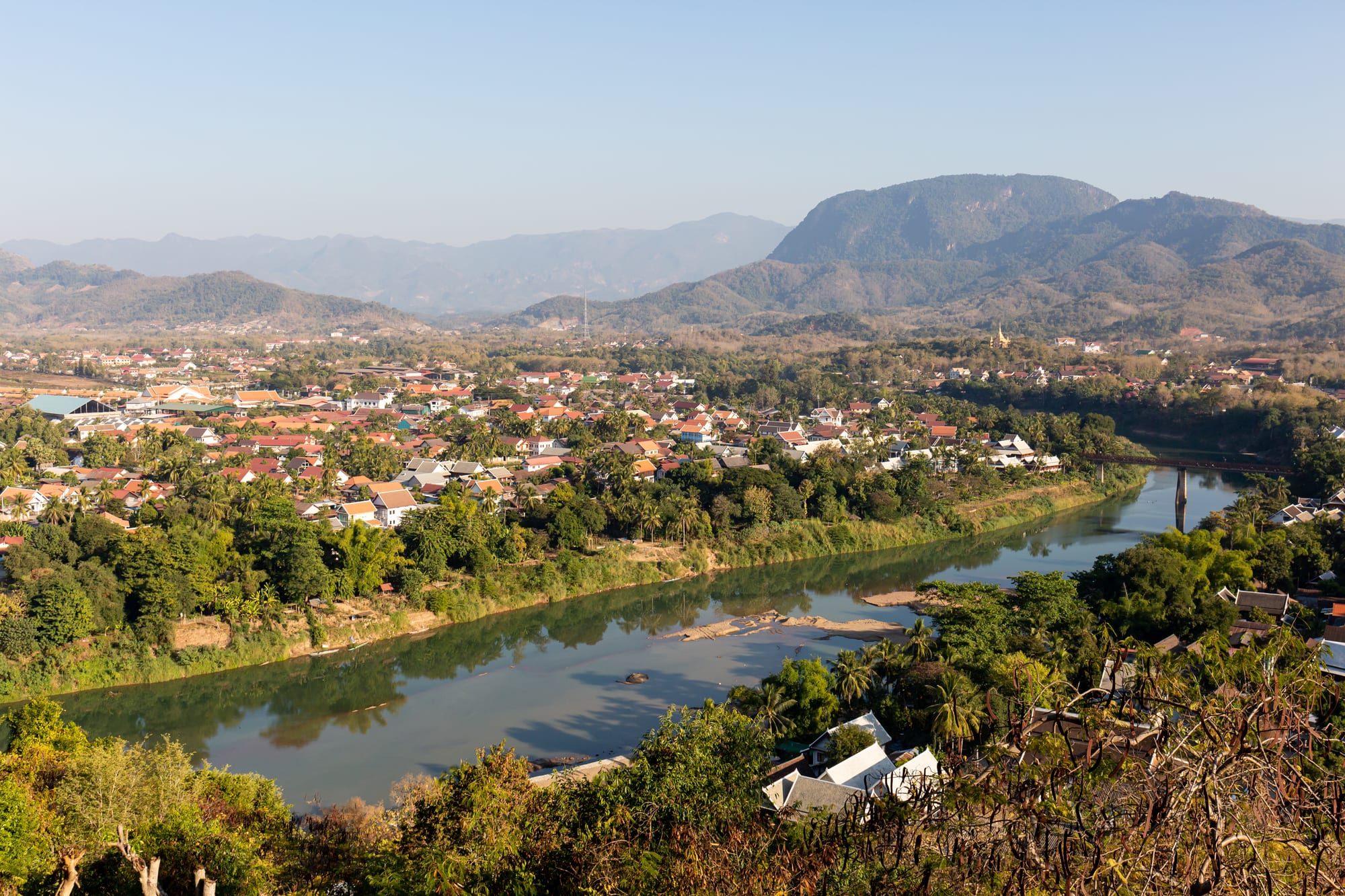 A small riverside town of temple roofs and old buildings seen from a hilltop, forested hills beyond