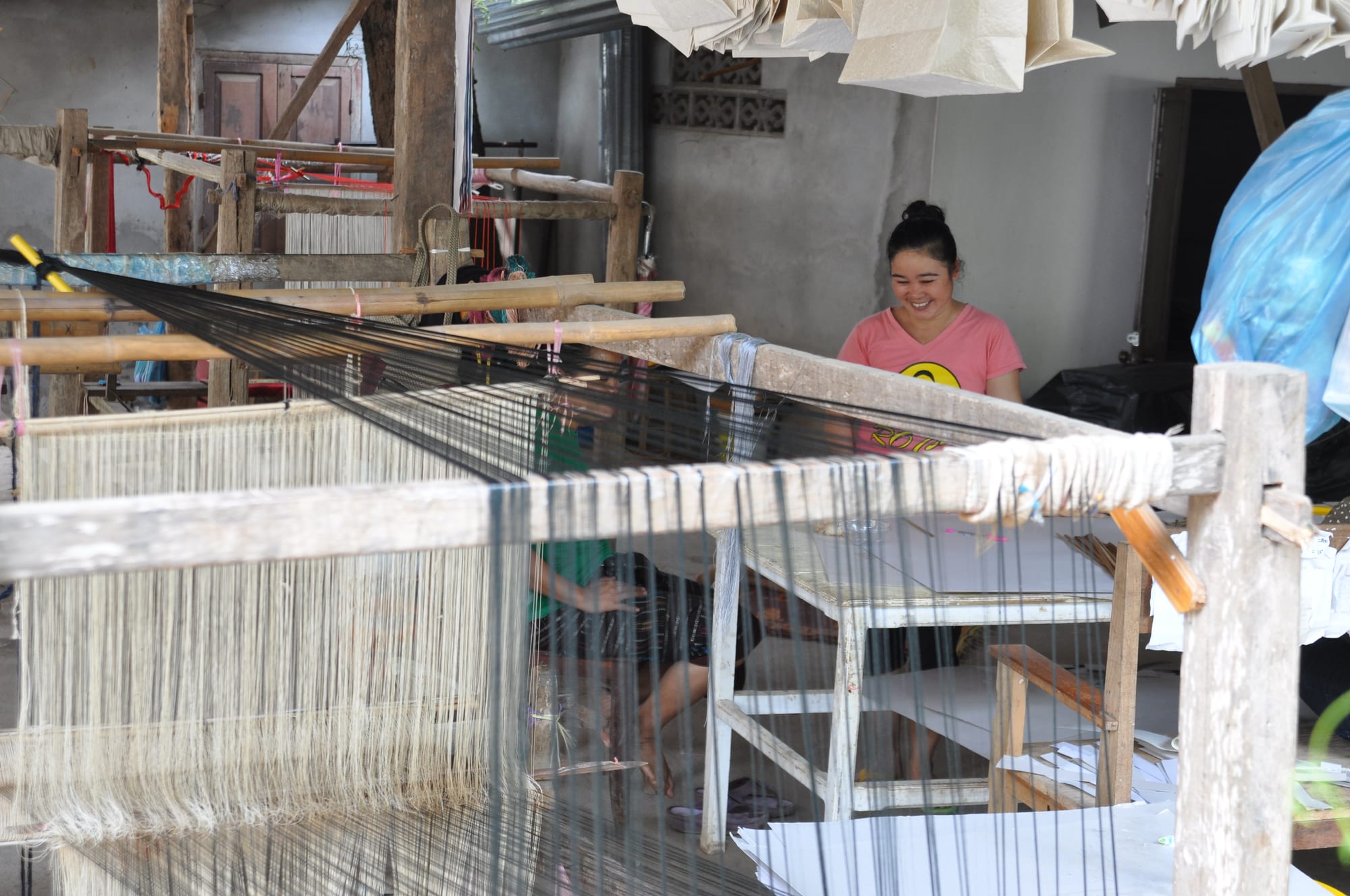 A smiling woman in a pink T-shirt sits behind a large wooden loom strung with long pale warp threads, with other wooden looms in the workshop behind her