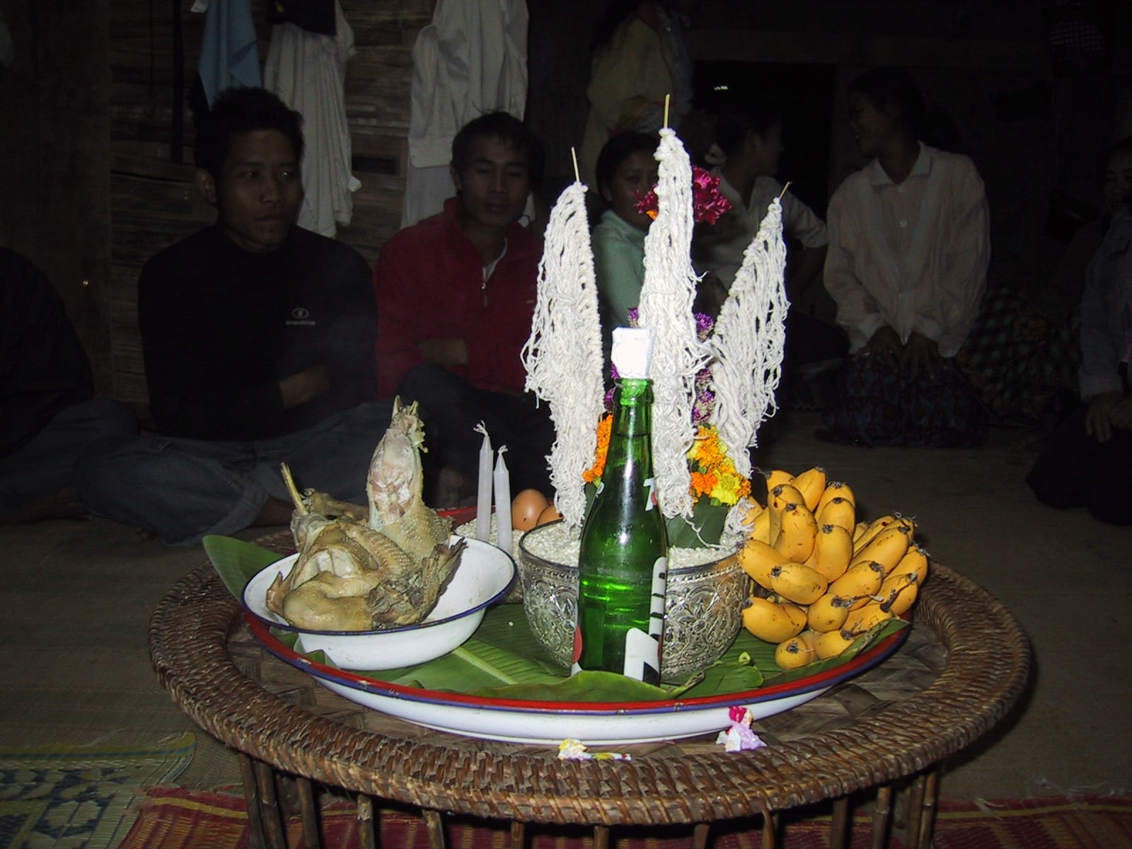 A rattan tray on a low table holding a centrepiece of rice and flowers hung with white cotton strings, beside a plate of cooked chicken, bananas, candles and a green bottle, with villagers seated on the floor behind
