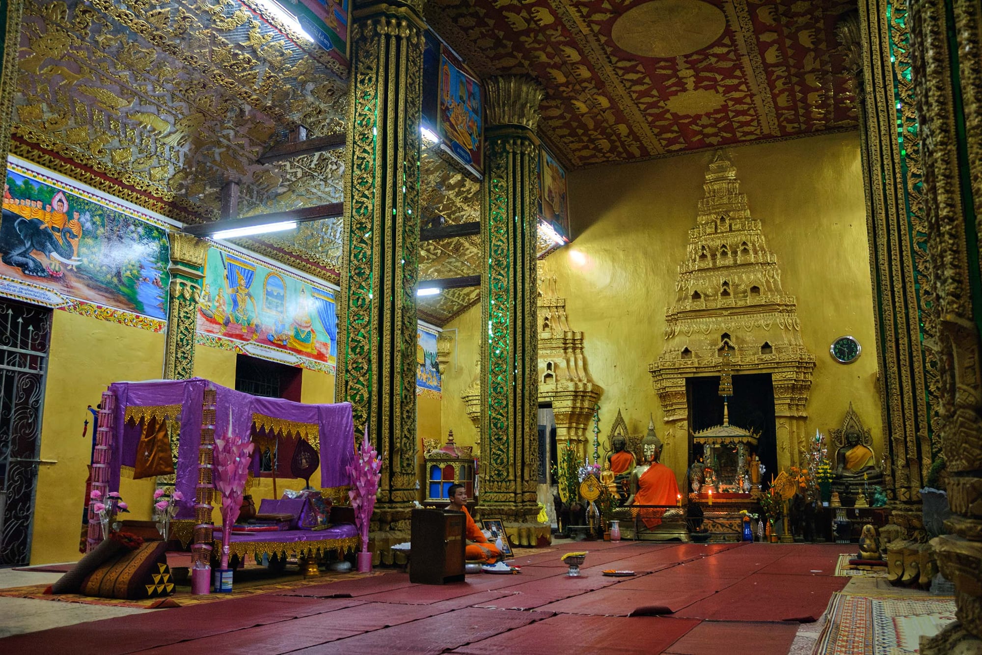 The interior of a Buddhist temple with gilded pillars, painted murals on yellow walls, a red floor and a tiered golden shrine at the far end, with a monk in an orange robe seated on the floor