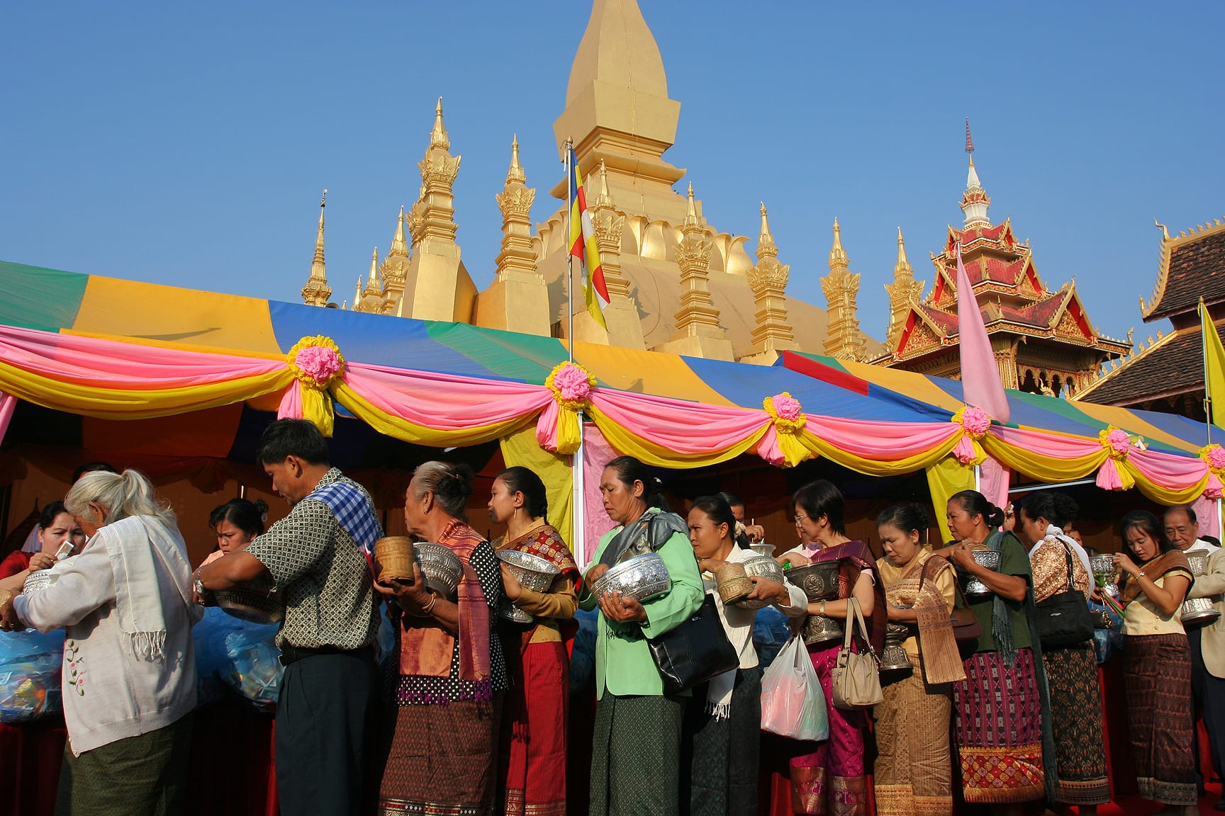 A line of women and men in patterned sinh skirts and sashes holding silver offering bowls beneath a pink and yellow awning, with a golden stupa rising behind them against a blue sky
