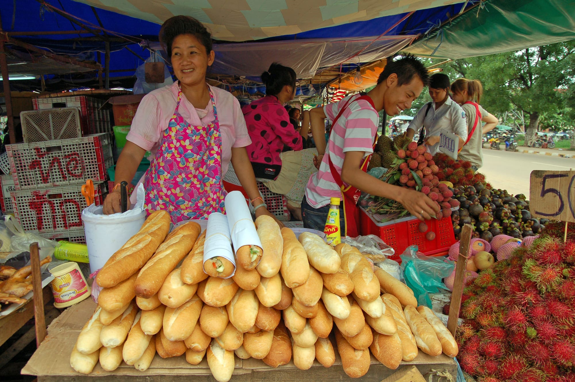 Market vendors at a stall piled with baguettes, rambutan, lychee, mangosteen and durian