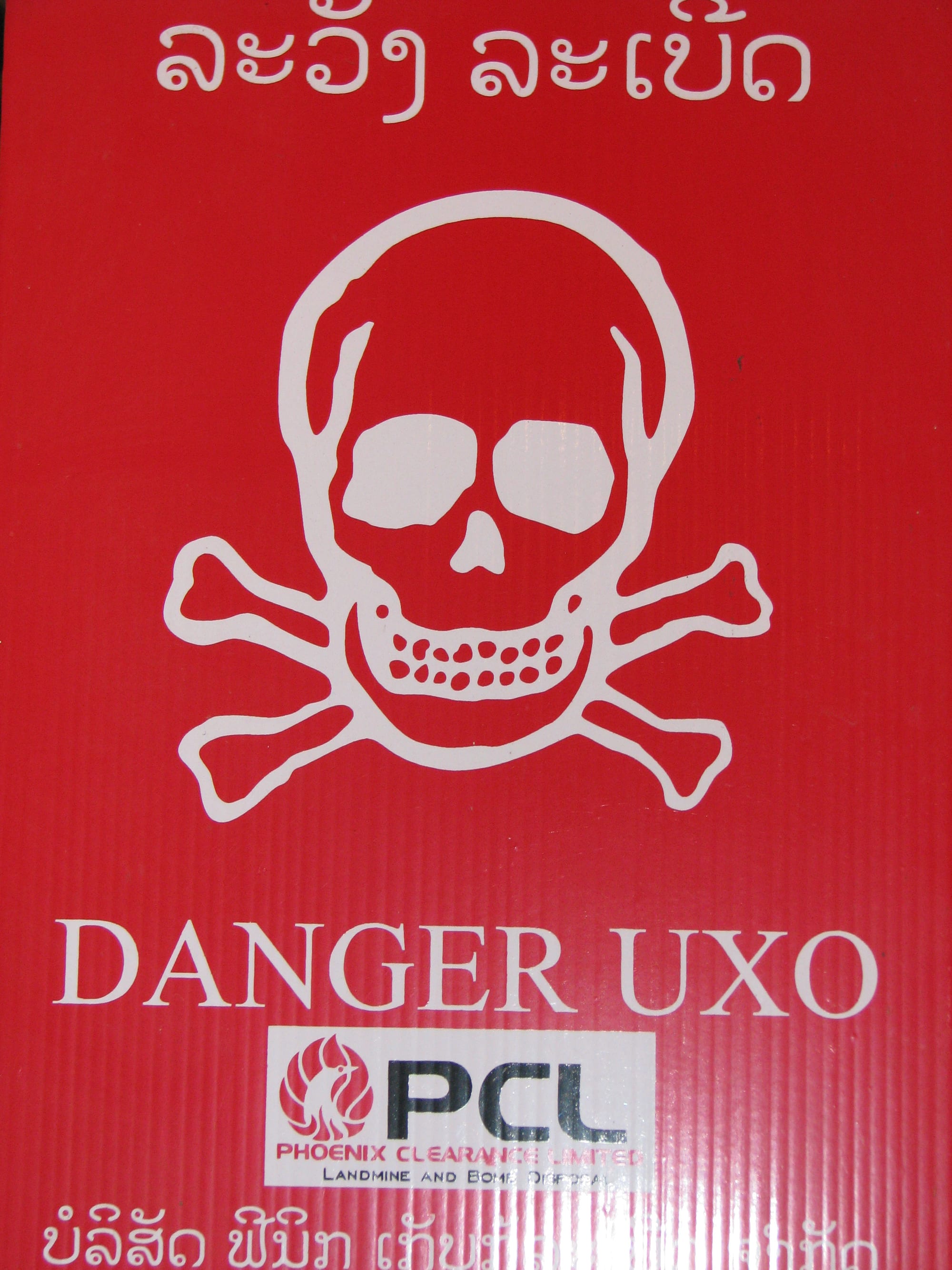 A hand-painted skull-and-crossbones sign warning of unexploded ordnance beside a rural path in Laos