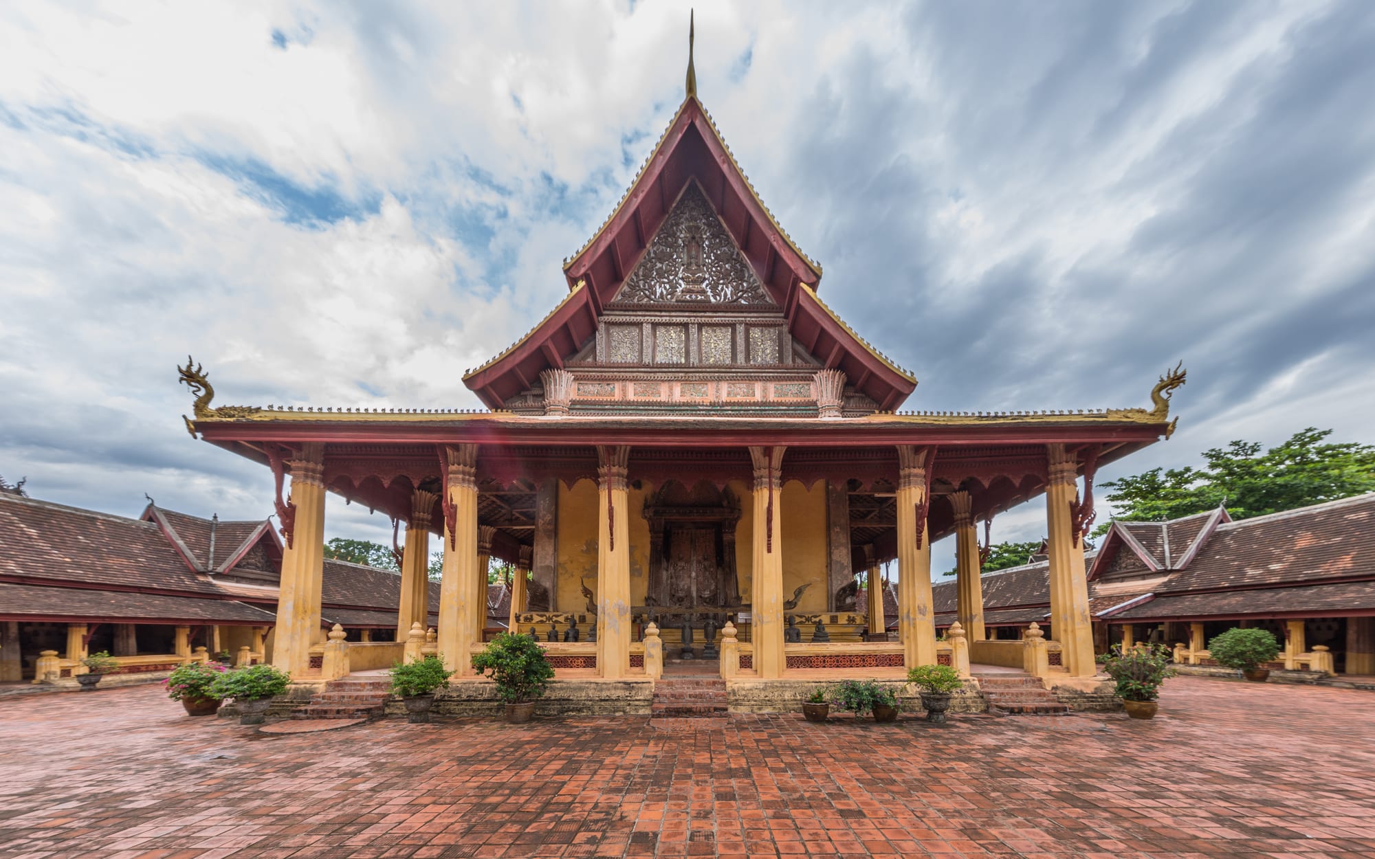 A Buddhist temple hall with a steep tiered red roof, carved gilded gable and a colonnaded veranda, standing in a brick-paved cloistered courtyard under heavy clouds