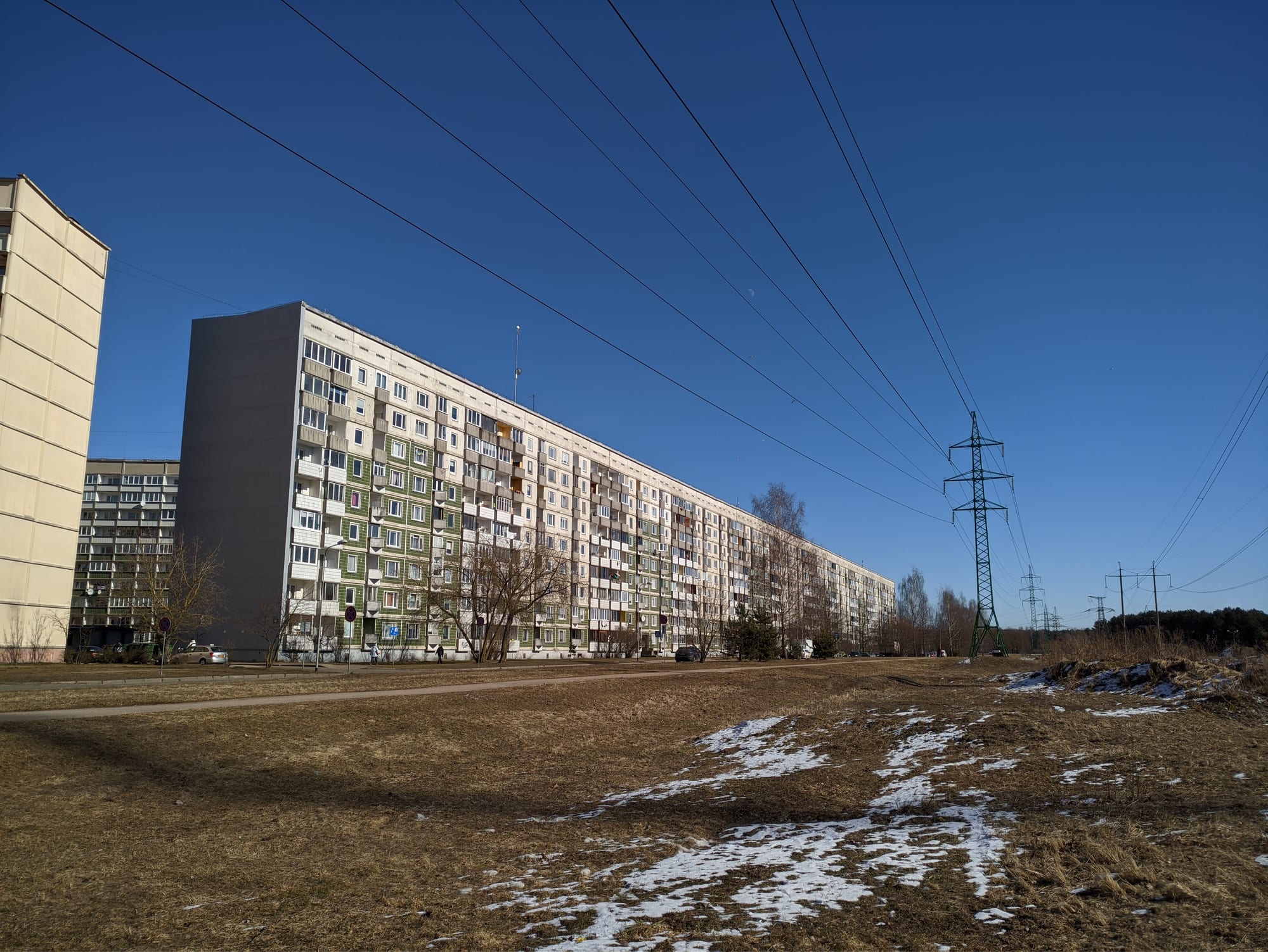 A long, multi-storey Soviet-era apartment block under a clear blue winter sky, with a smaller building and electricity pylons nearby