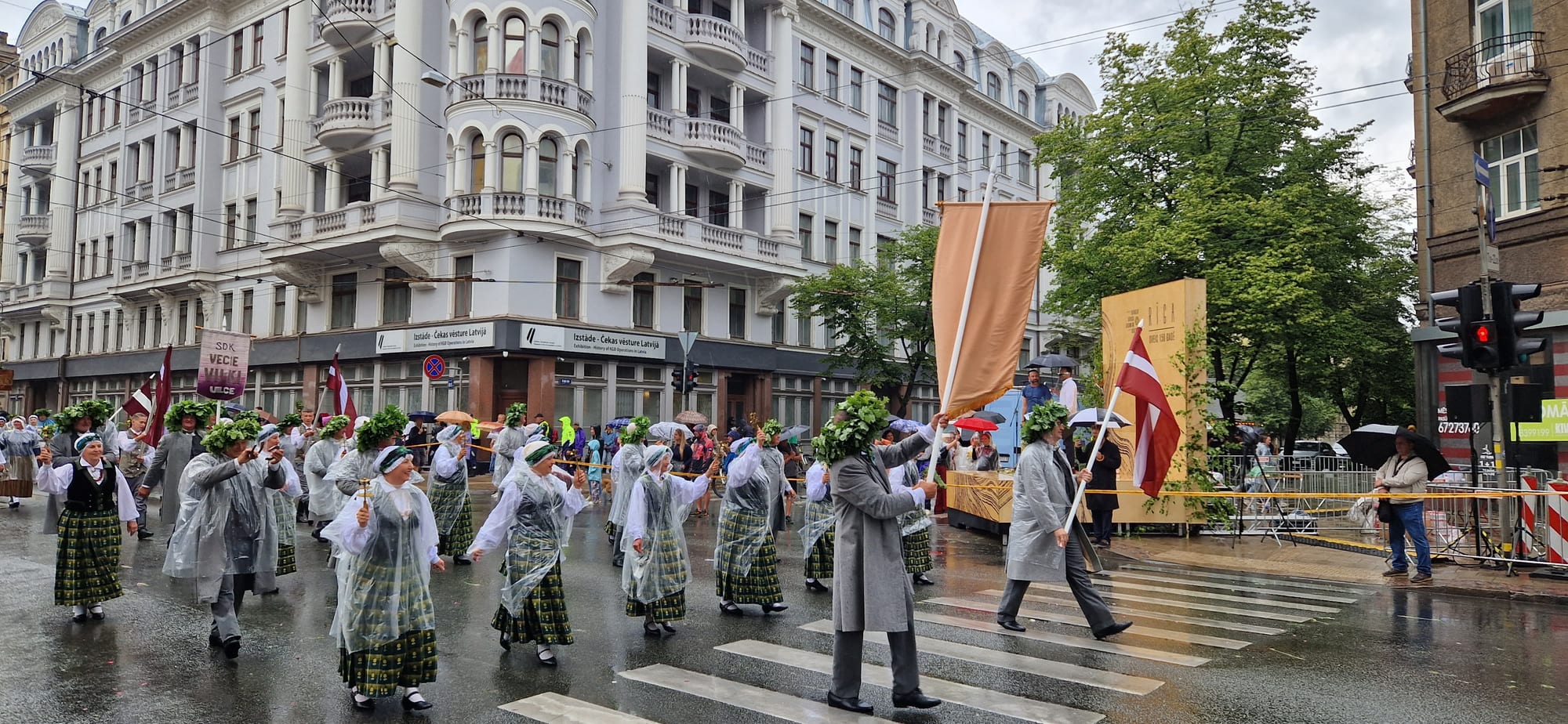 A procession of Latvians in traditional folk dress, wearing green leaf wreaths and carrying flags, marching through a city street in the rain