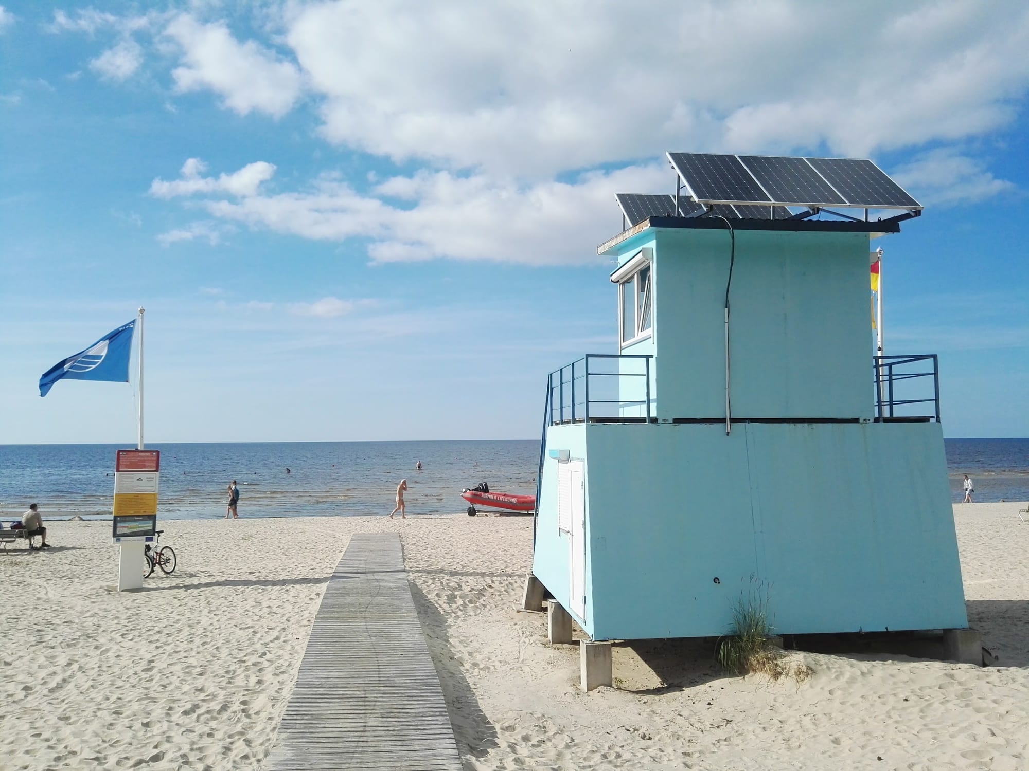 A wide white-sand beach with wooden groynes stretching to calm sea, pine trees behind