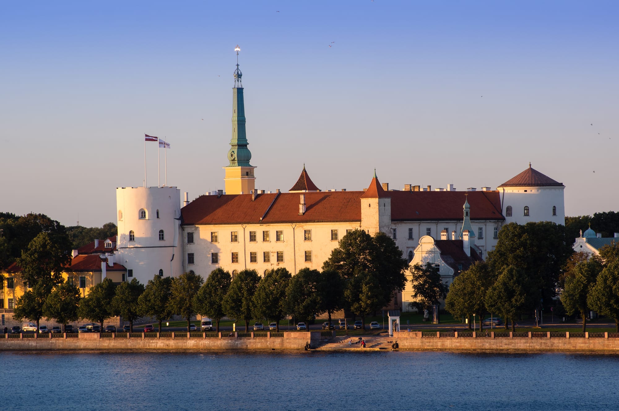 A long cream-coloured castle with round white corner towers, red roofs and a slender green spire, standing behind a line of trees on a river embankment in evening light