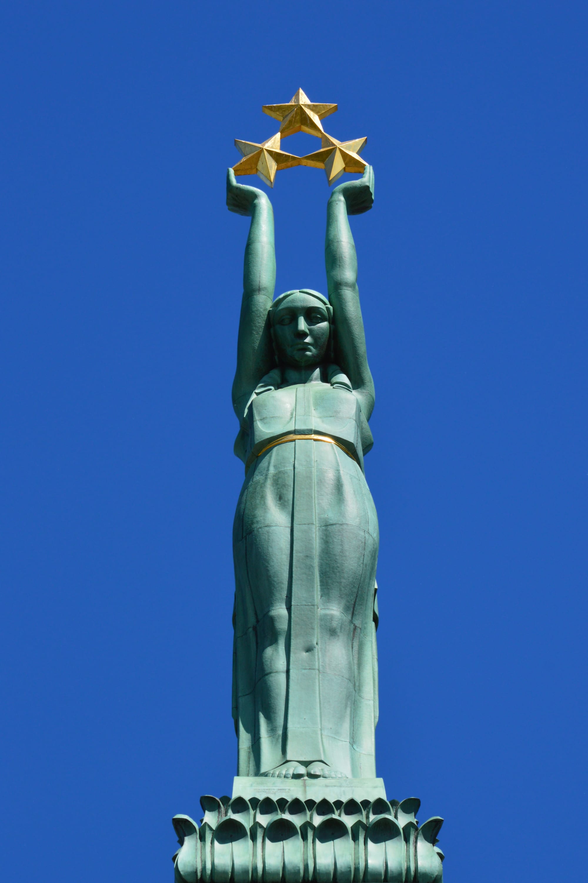 A tall stone column topped by a bronze female figure holding up three gold stars, in Riga