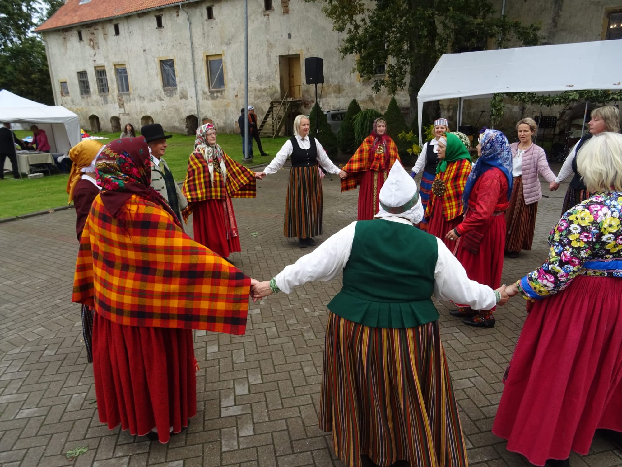 Women in striped and red skirts, bright checked shawls and headscarves dance in a circle holding hands in a cobbled courtyard in front of an old whitewashed building
